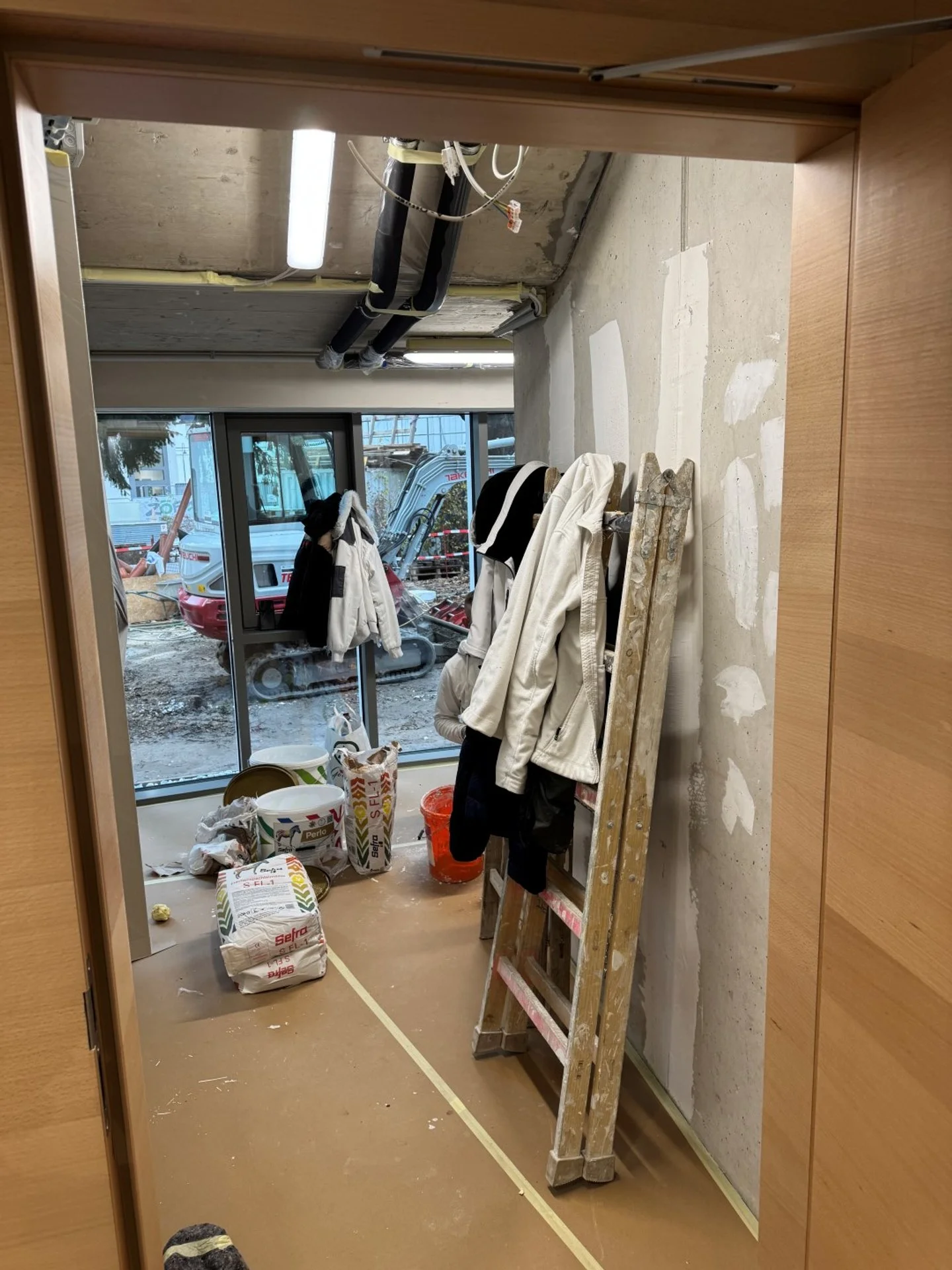 Innenaufnahme eines Raumes im Bau oder Renovierungsprozess mit großer Glasschiebetür, Baumaterialien, eine Leiter mit Kleidung und Bauutensilien, draußen Bauarbeiten sichtbar. Von bmk-projekt umgesetzt in Wien.