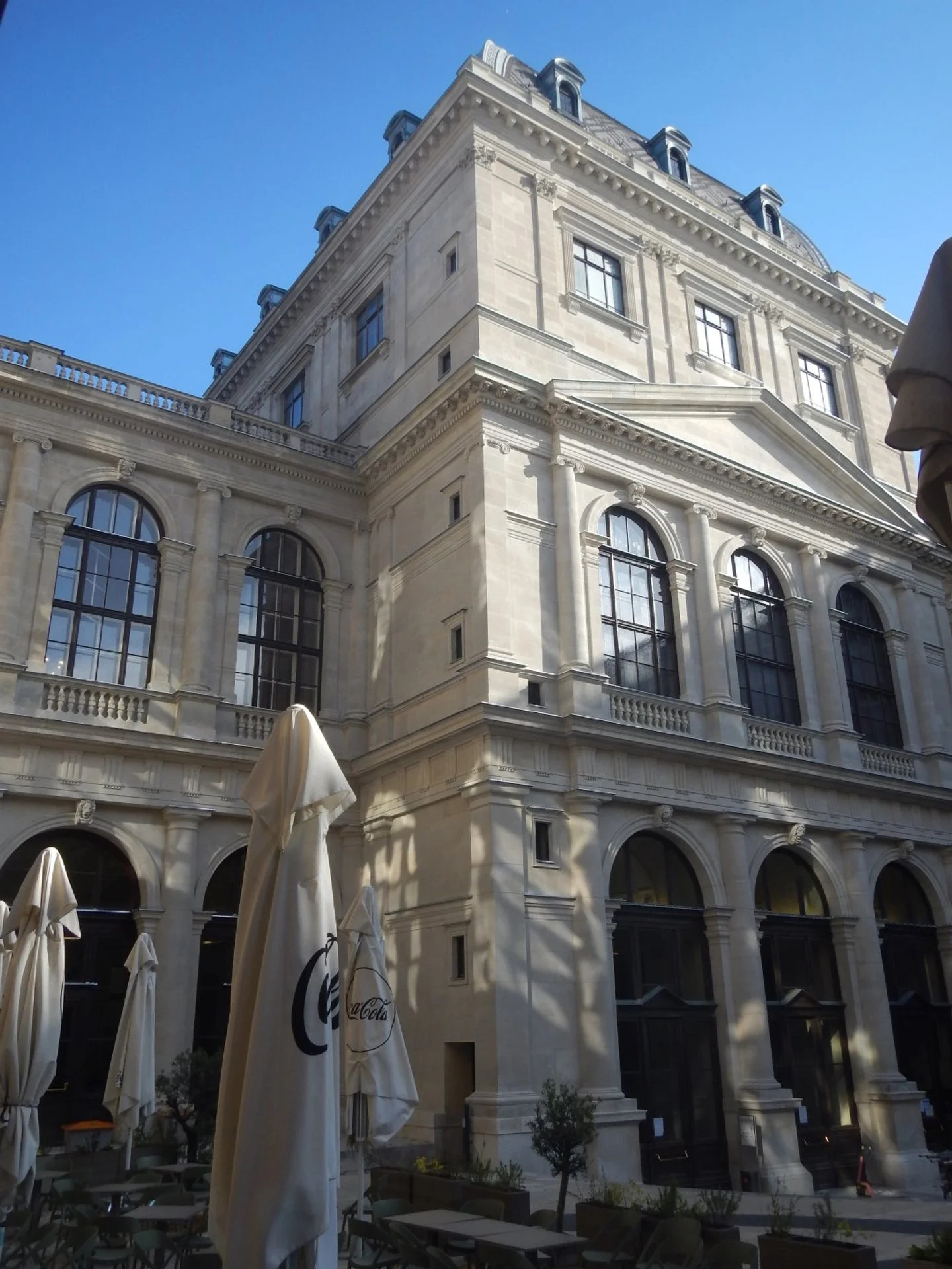 Ein historisches, mehrstöckiges Gebäude mit großen Fenstern, Verzierungen und einem Dach mit kleinen Terrassen im Vordergrund befinden sich Sonnenschirme.