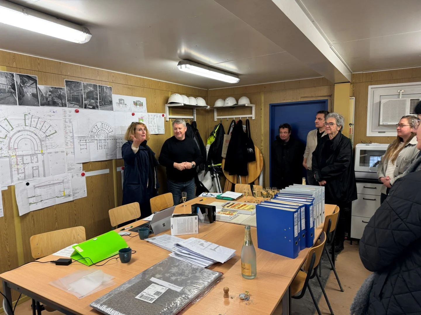 Ein Gruppenfoto während eines Besprechungsraums, mit mehreren Personen, die auf eine Frau zuhören, die spricht. Tische mit Unterlagen, Ordnern und Getränken im Raum, an der Wand Baupläne und Fotos.