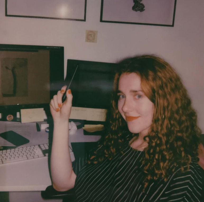 A woman with curly red hair sitting at a desk in front of a computer, holding a stylus, with a drawing of a dress on the computer screen.