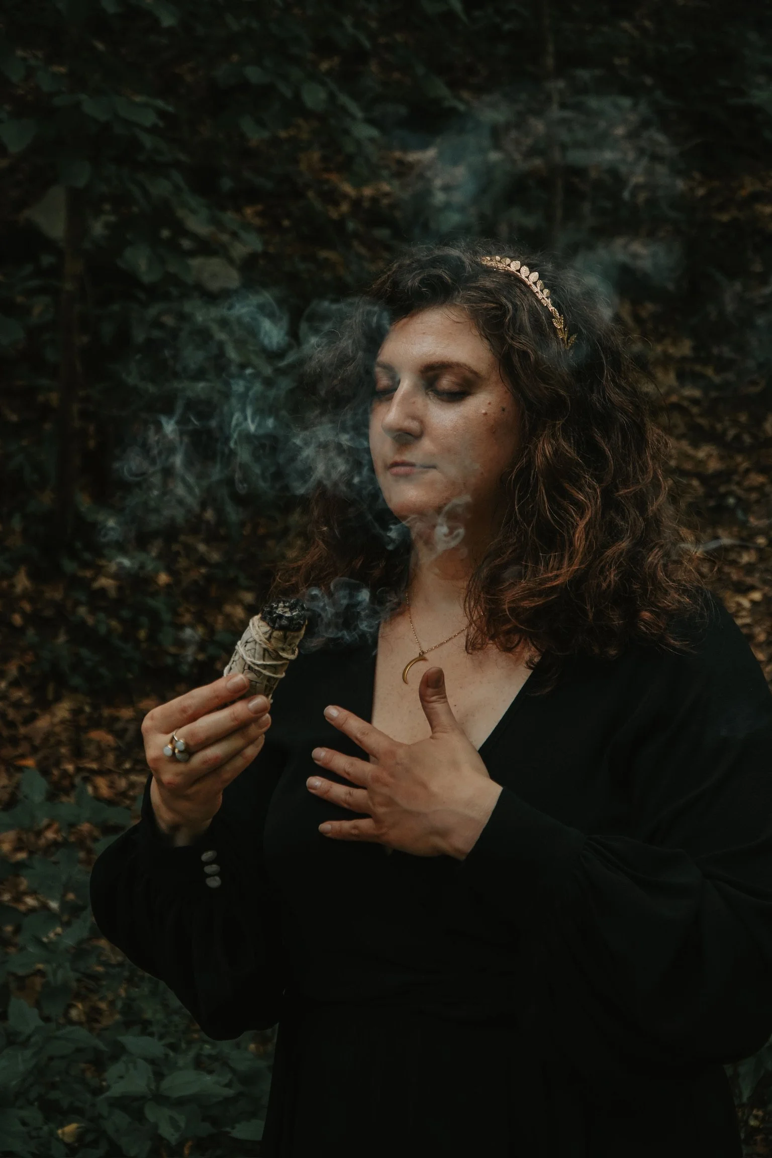 A woman with curly hair, wearing a black dress, a gold headband, and a crescent moon necklace, stands outdoors in a forested area, holding a lit sage bundle emitting smoke, with her eyes closed.
