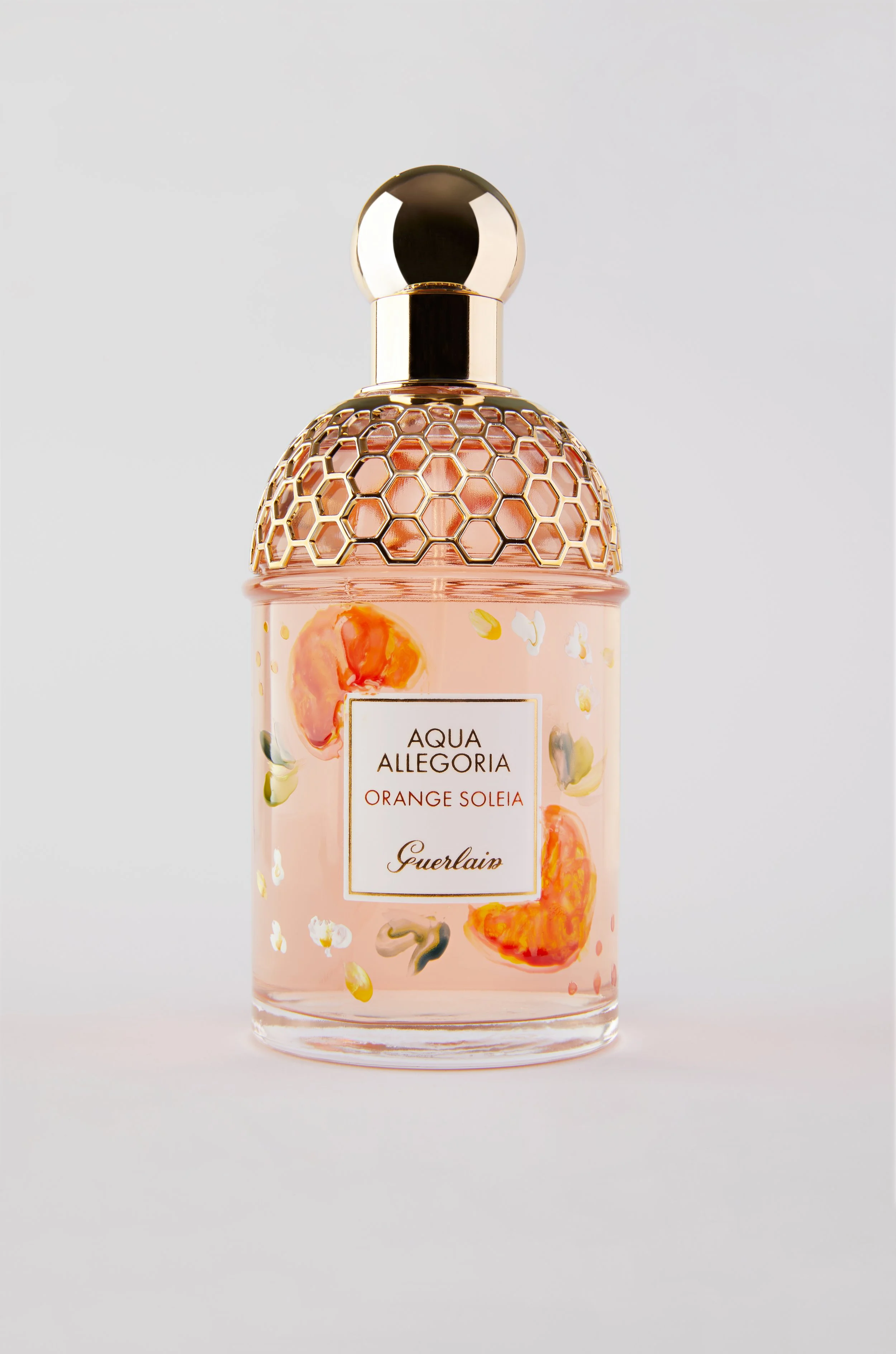 A perfume bottle with a clear glass body, gold cap, and decorative honeycomb pattern, labeled 'Aqua Allegoria Orange Soleil Guerlain' with a peach and orange flower print around the label.