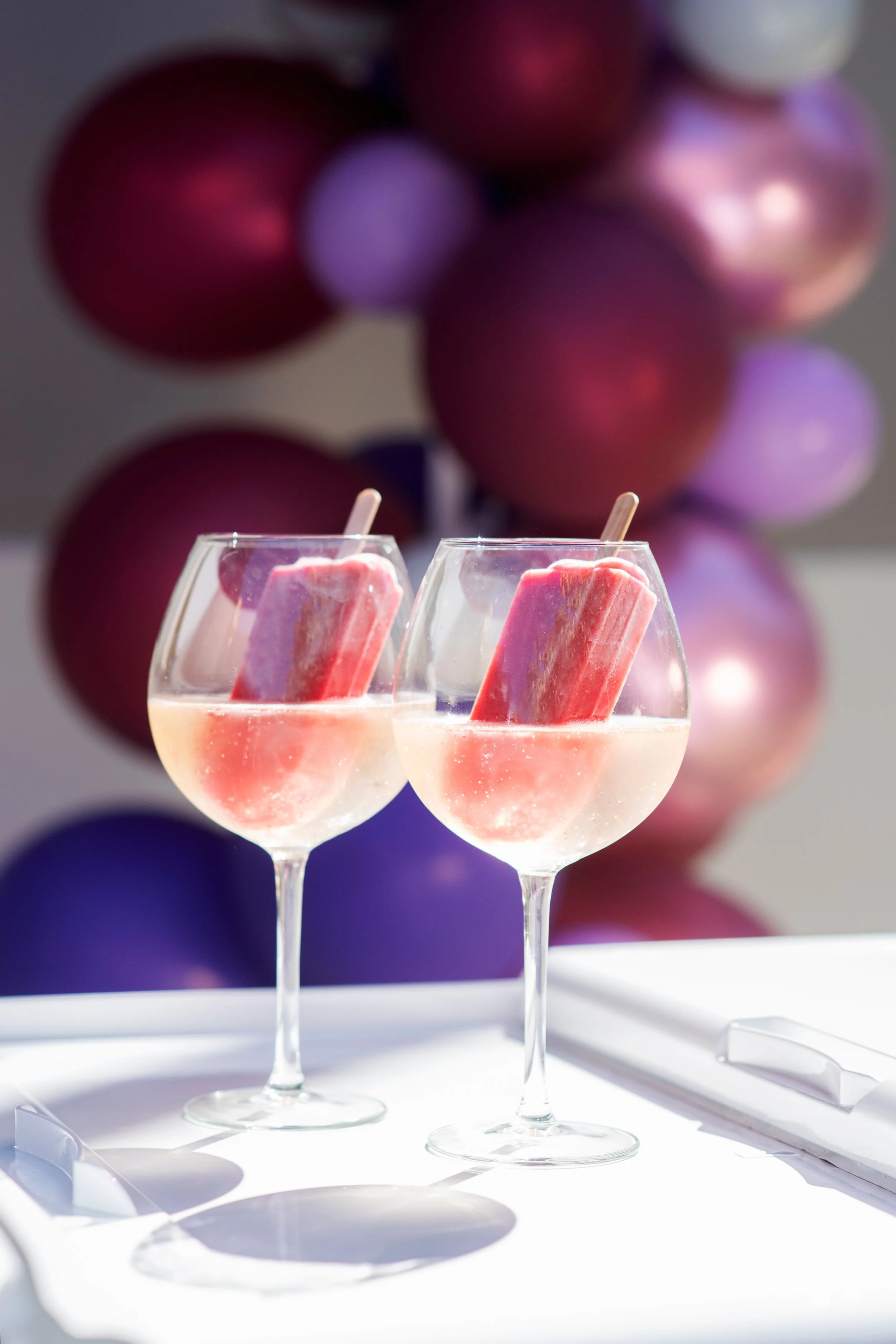 Two elegant wine glasses filled with a pale pink beverage, each with a pink popsicle inside, on a white table with colorful balloons in the background.