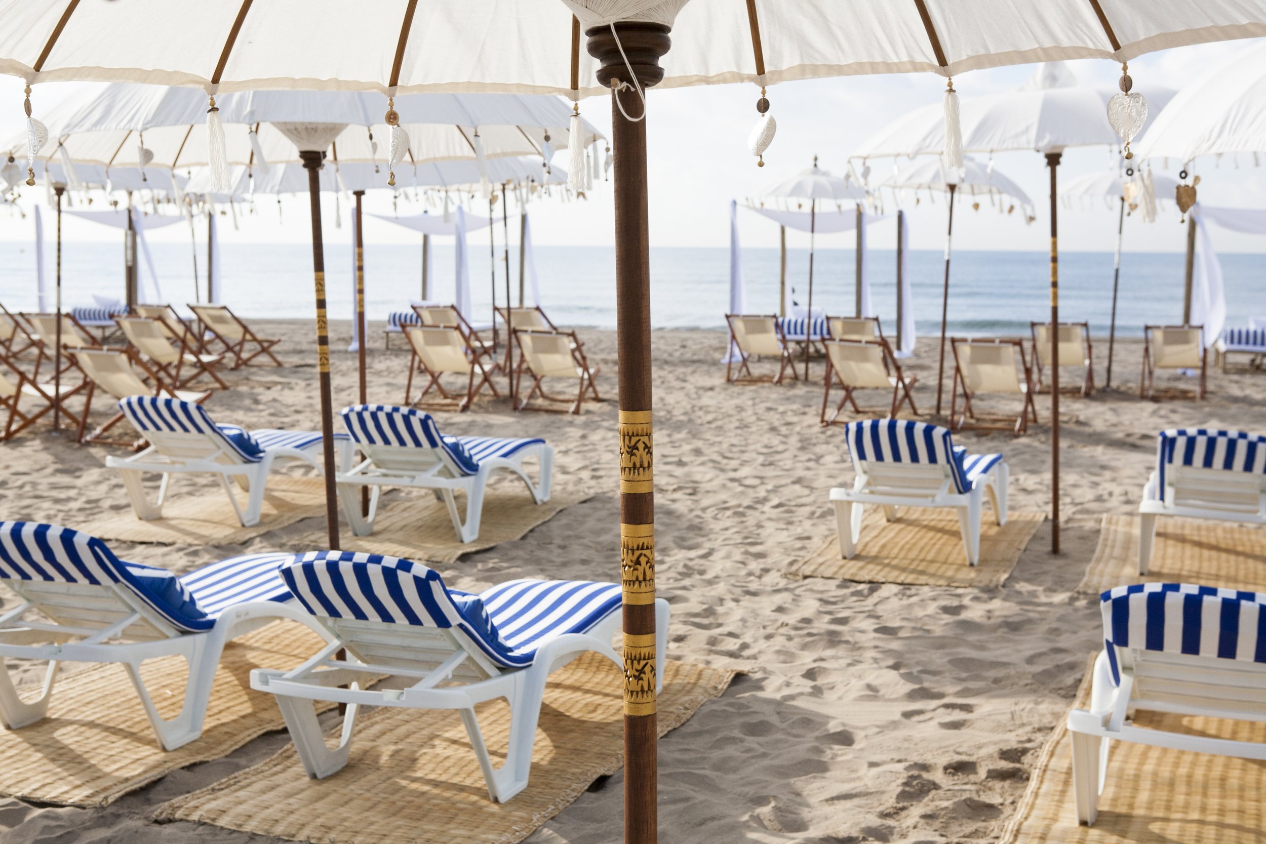 Empty beach with white umbrellas, chairs with blue and white striped cushions, and sandy ground leading to the ocean.