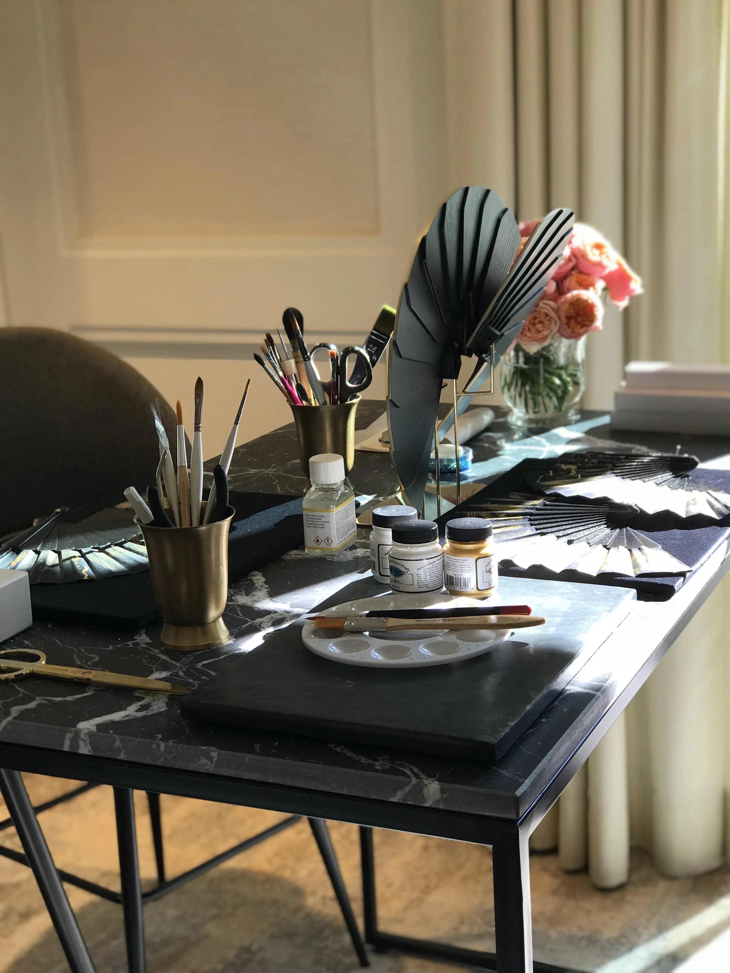 A table with painting supplies, brushes, paint bottles, and decorative fans, with a vase of pink roses in the background.