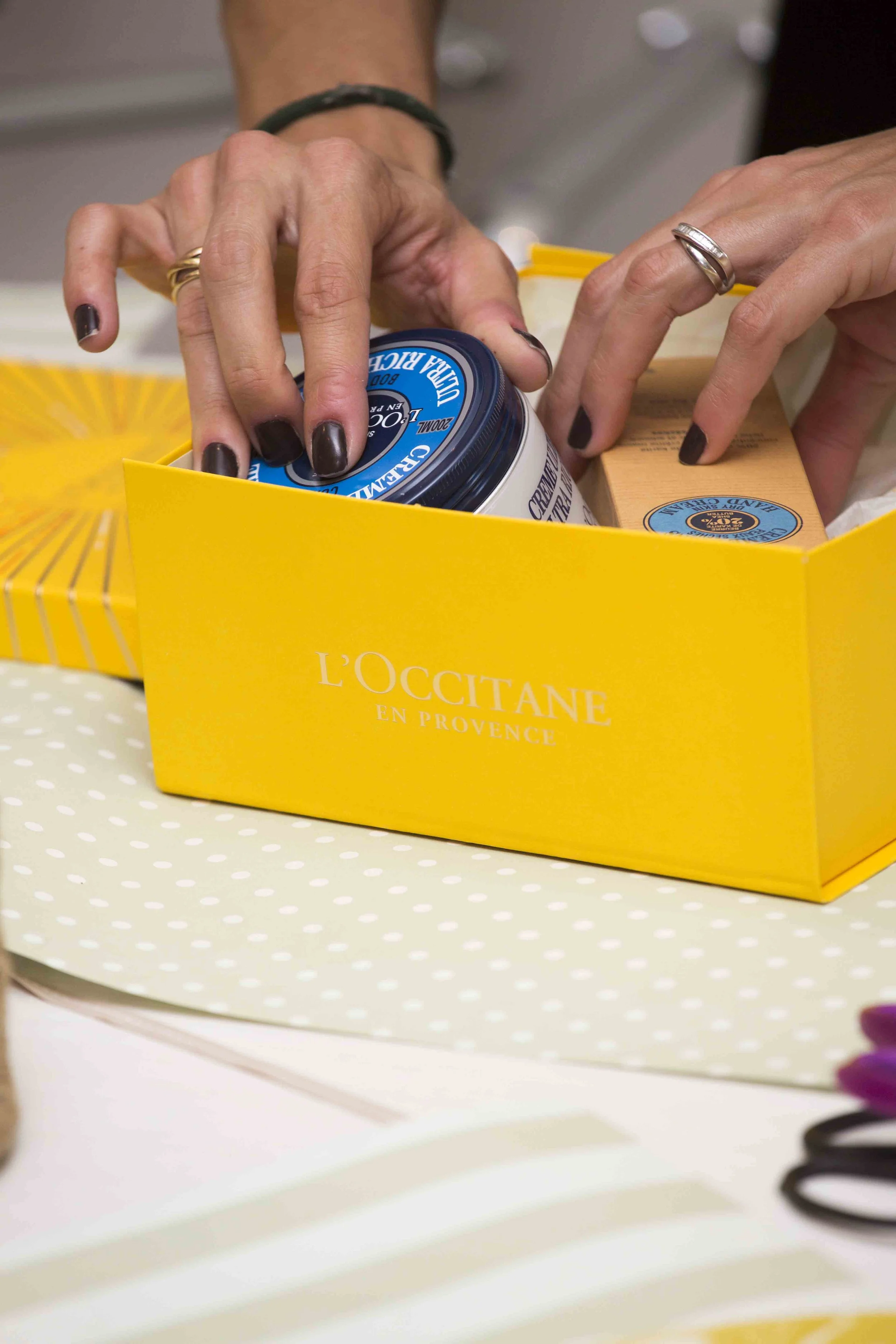 Hands organizing skincare products in a yellow L'Occitane gift box, including a cream jar and soap bars.