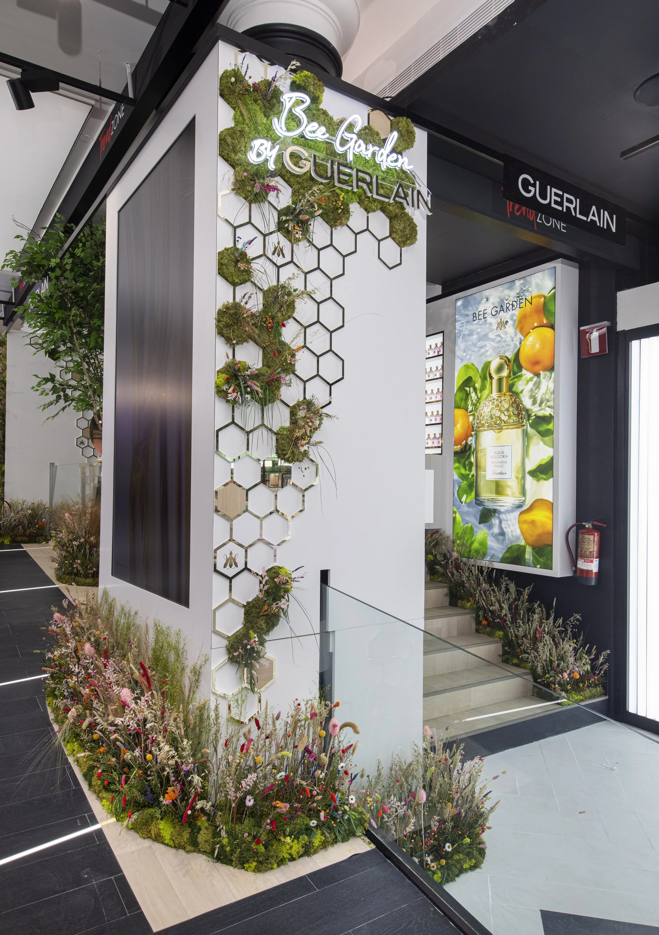 Interior shopping mall display for Bee Garden by Guerlain, featuring a floral-themed wall with honeycomb design and a large digital advertisement for a perfume surrounded by flowers.