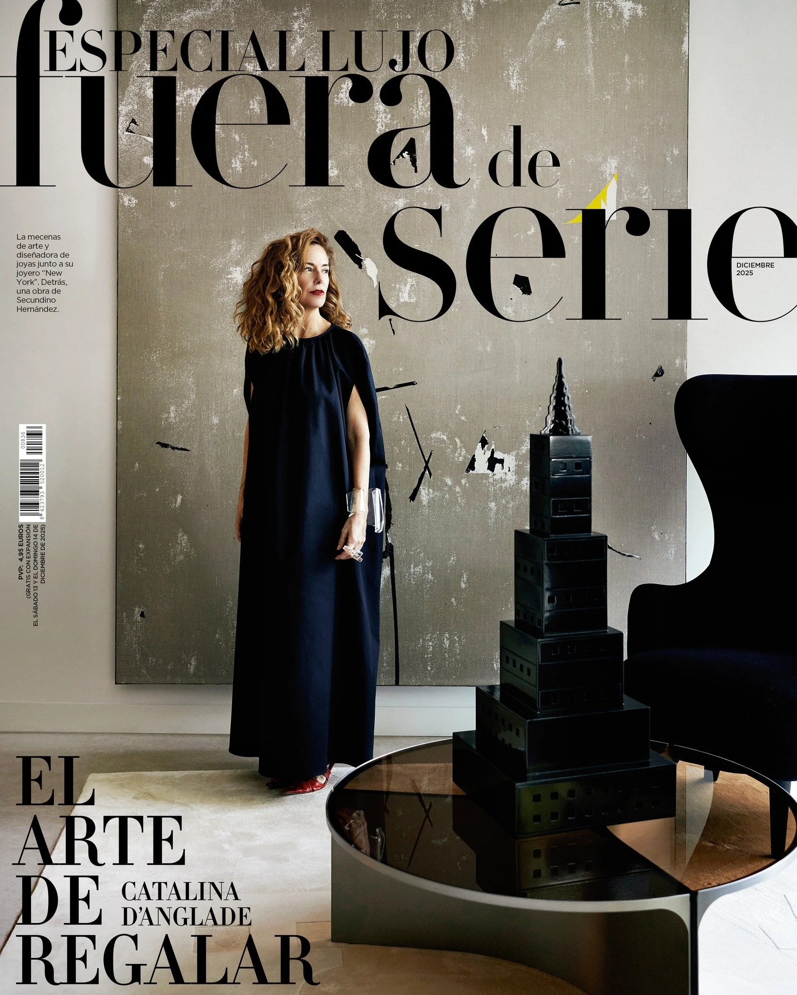 A woman with curly red hair dressed in a long black gown standing indoors next to a black armchair and a small round glass top table with a black architectural model resembling a high-rise building. Behind her, there is a textured wall with large black text and a smaller paragraph in Spanish. The woman is holding a glass and looking to her left.