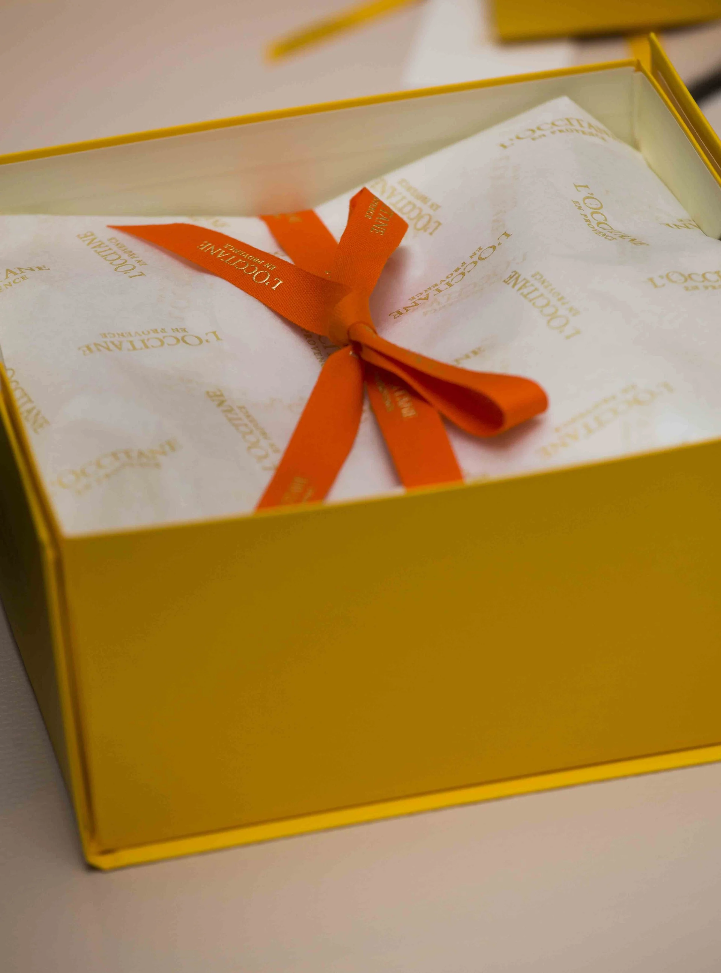 A yellow box with tissue paper inside, tied with an orange ribbon that has 'L'OCCITANE EN PROVENCE' printed repeatedly.