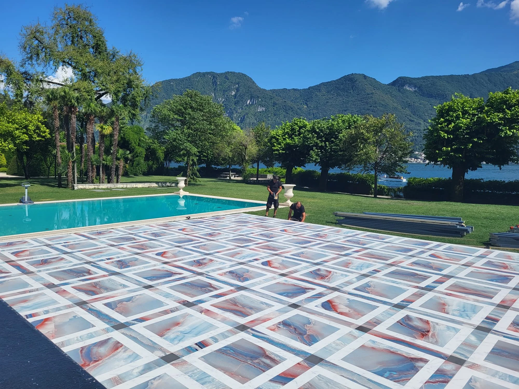 Persone che installano o sistemano piastrelle decorative vicino a una piscina all'aperto con vista montagna e alberi verdi, cielo blu e acqua sullo sfondo.