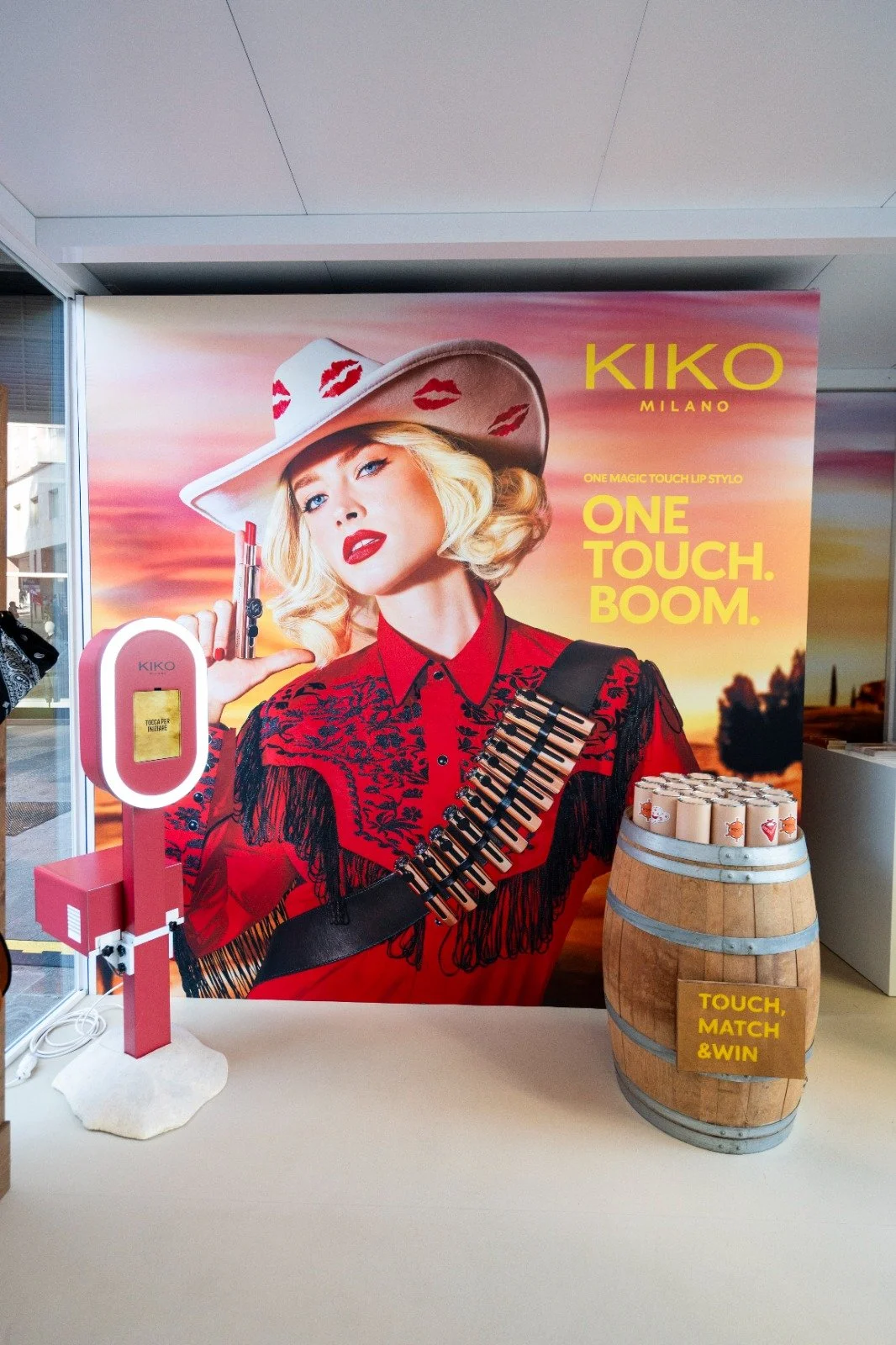 Poster pubblicitario di KIKO Milano con un'immagine di una donna con cappello bianco decorato con baci rossi, vestita con una camicia rossa e una fascia con tasti di pianoforte come stretta sulla spalla. C'è anche una cabina telefonica rosa e un bari