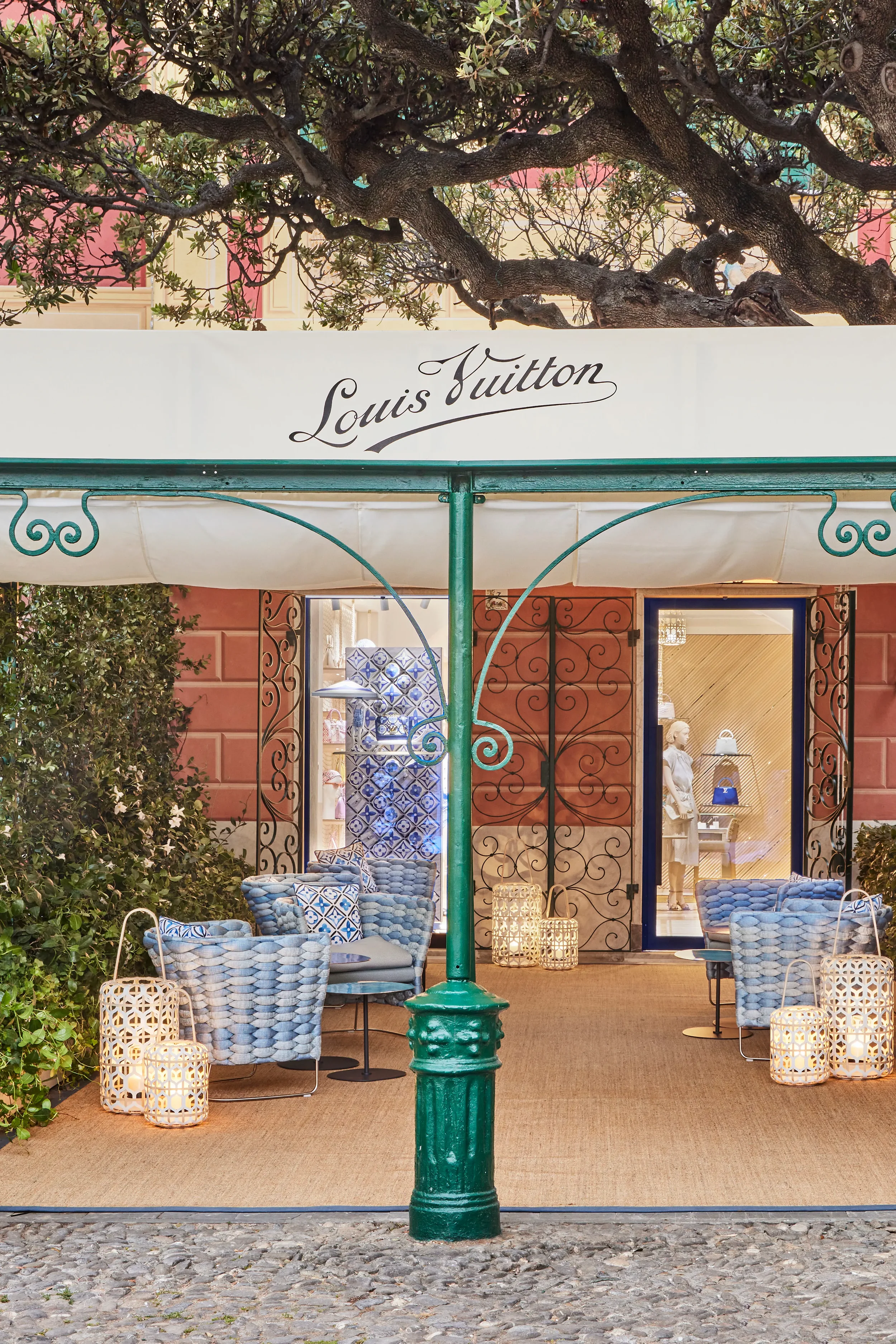 Esterno di un negozio di Louis Vuitton con arredamento elegante e decorazioni luminose, sedie e lanterne, siepi verdi e albero sopra l'ingresso.