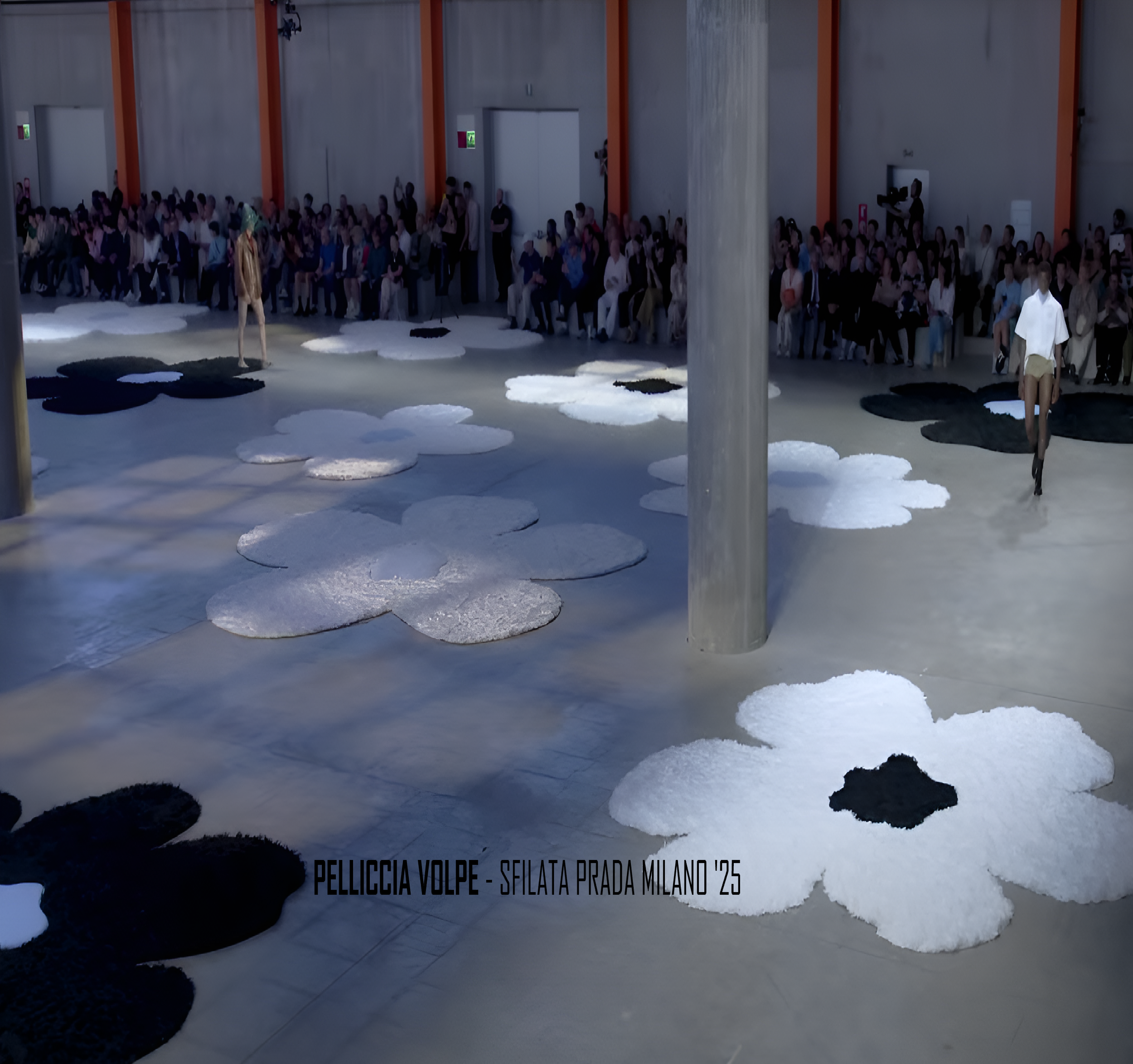 Mostra di moda con modelli che indossano abbigliamento su un palco decorato con tappeti a forma di fiori, audience seduta sullo sfondo in un grande spazio industriale. L'installazione si chiama 'Pelliccia Volpe' presentata alla sfilata Prada Milano 2