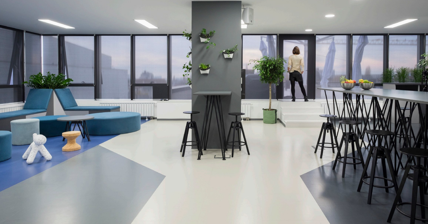 Interno di un moderno spazio ufficio o lounge con grandi finestre, piante verdi, sedute colorata, banconi alti e una persona che guarda fuori dalla finestra.