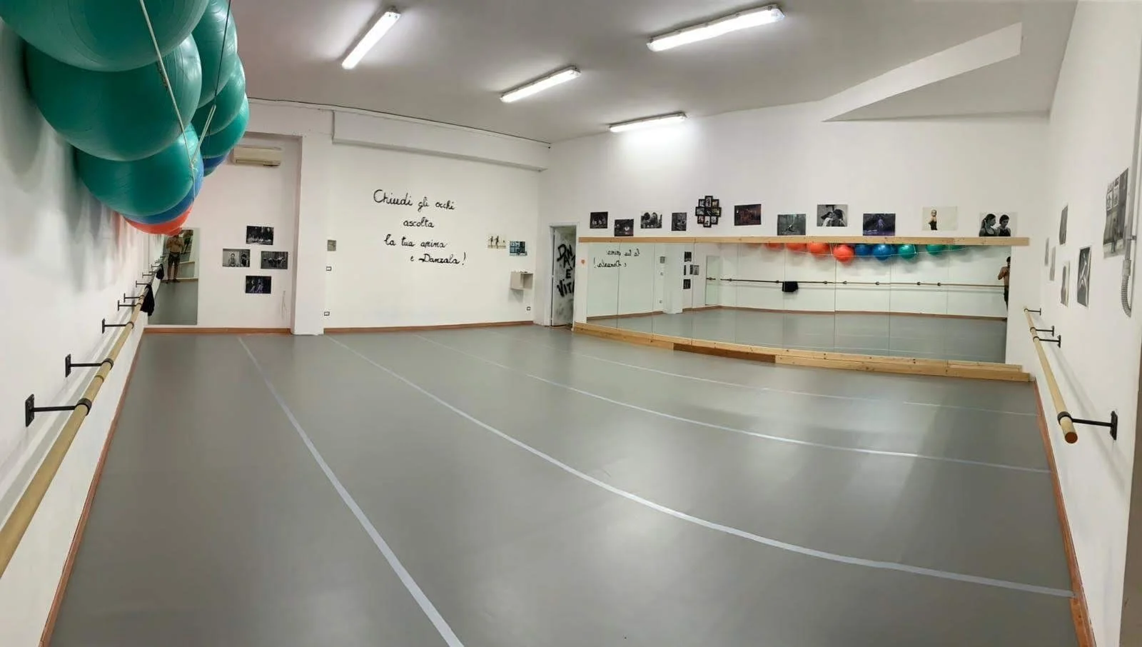 Studio di danza vuoto con pareti bianche, specchi a tutta altezza, rulli di bastoni per esercizi di danza, palloni di fitness colorati appesi, e foto alle pareti.