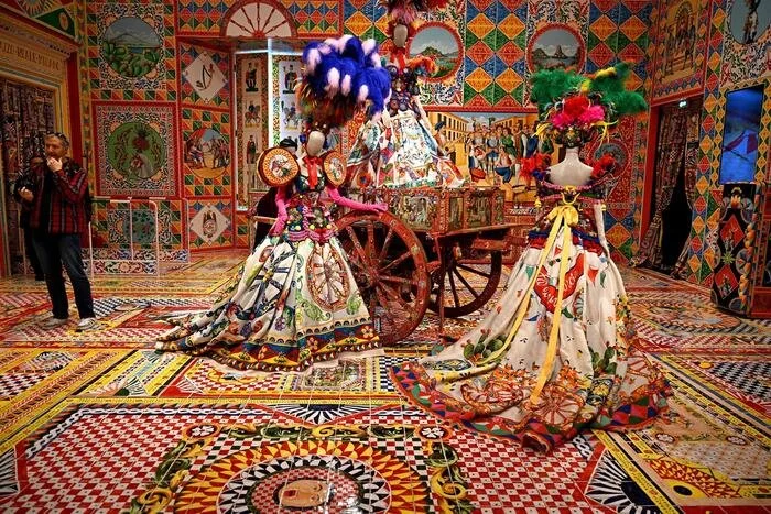 L'immagine mostra una stanza decorata con motivi colorati e geometrici sulle pareti e sul pavimento. Al centro, ci sono due manichini vestiti con abiti tradizionali ricchi di colori e ornamenti, e una carrozza decorata con fiori e tessuti vari.