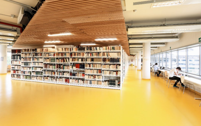 Libreria moderna con scaffali pieni di libri, soffitto in legno curvo, pavimento giallo, zona di lettura con grandi finestre e persone che leggono.