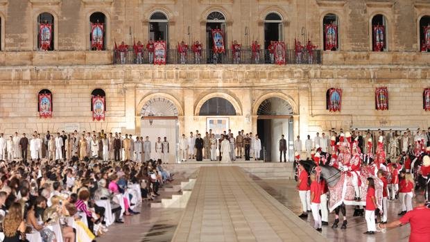 Una cerimonia con persone in abiti tradizionali davanti a un palazzo storico, con un pubblico seduto e un corteo di cavalli decorati.