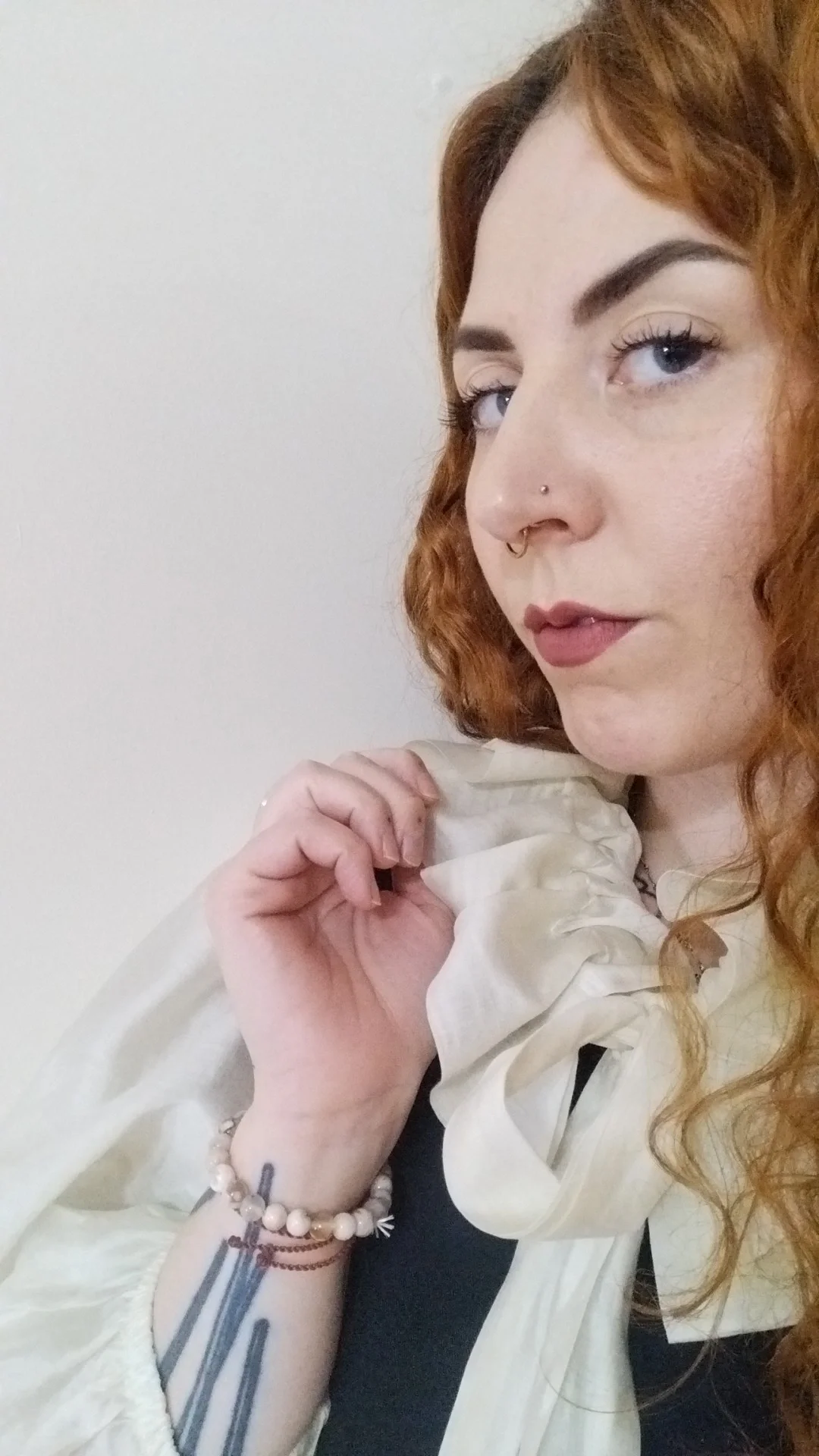 Close-up of a woman with curly red hair, blue eyes, pierced nose, and wearing a cream-colored top with a large bow, holding the collar with one hand, and wearing bracelets.