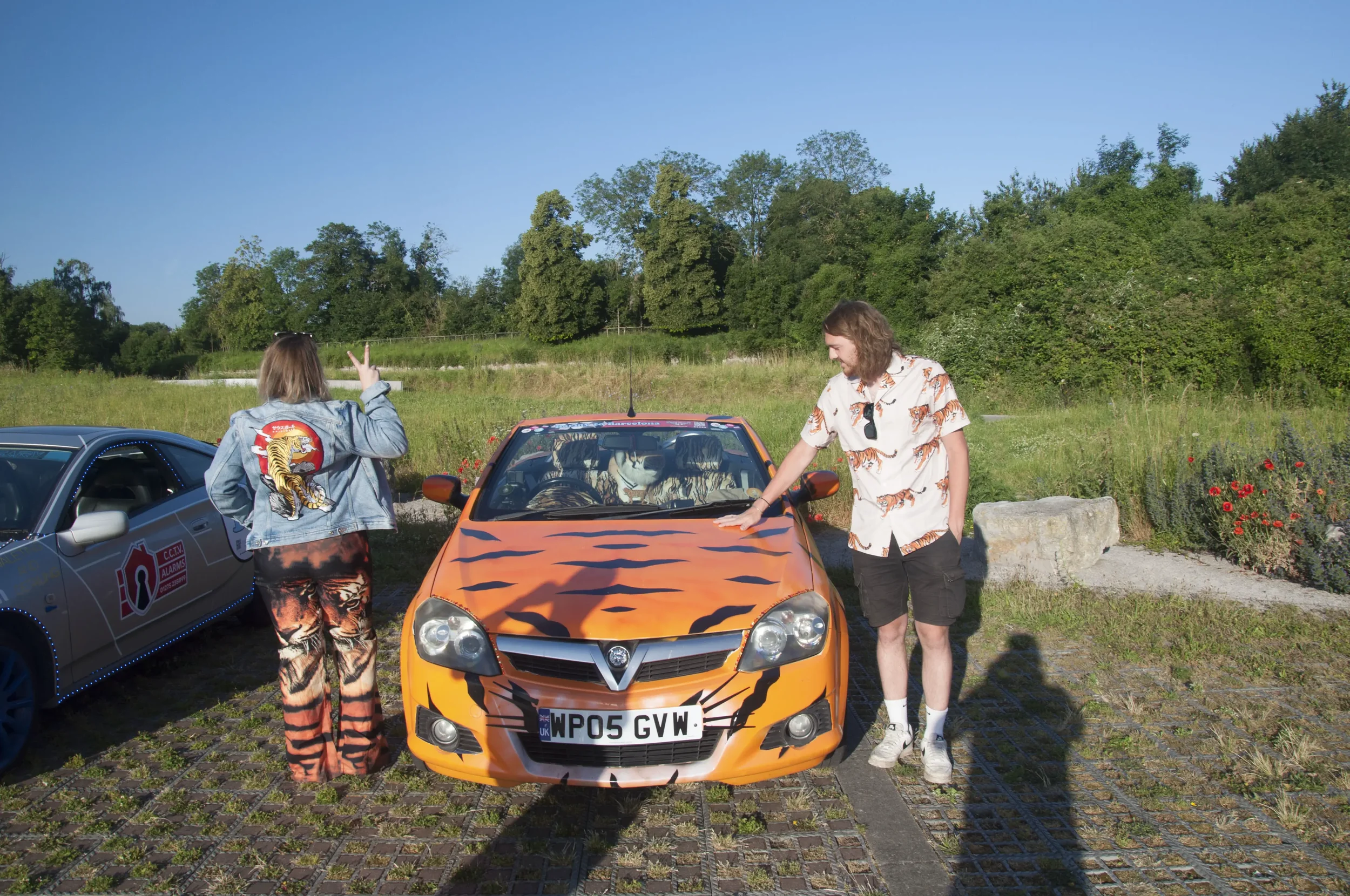 Two people standing next to a bright orange car with tiger stripe design on the hood in an outdoor parking area, with grassy field and trees in the background.
