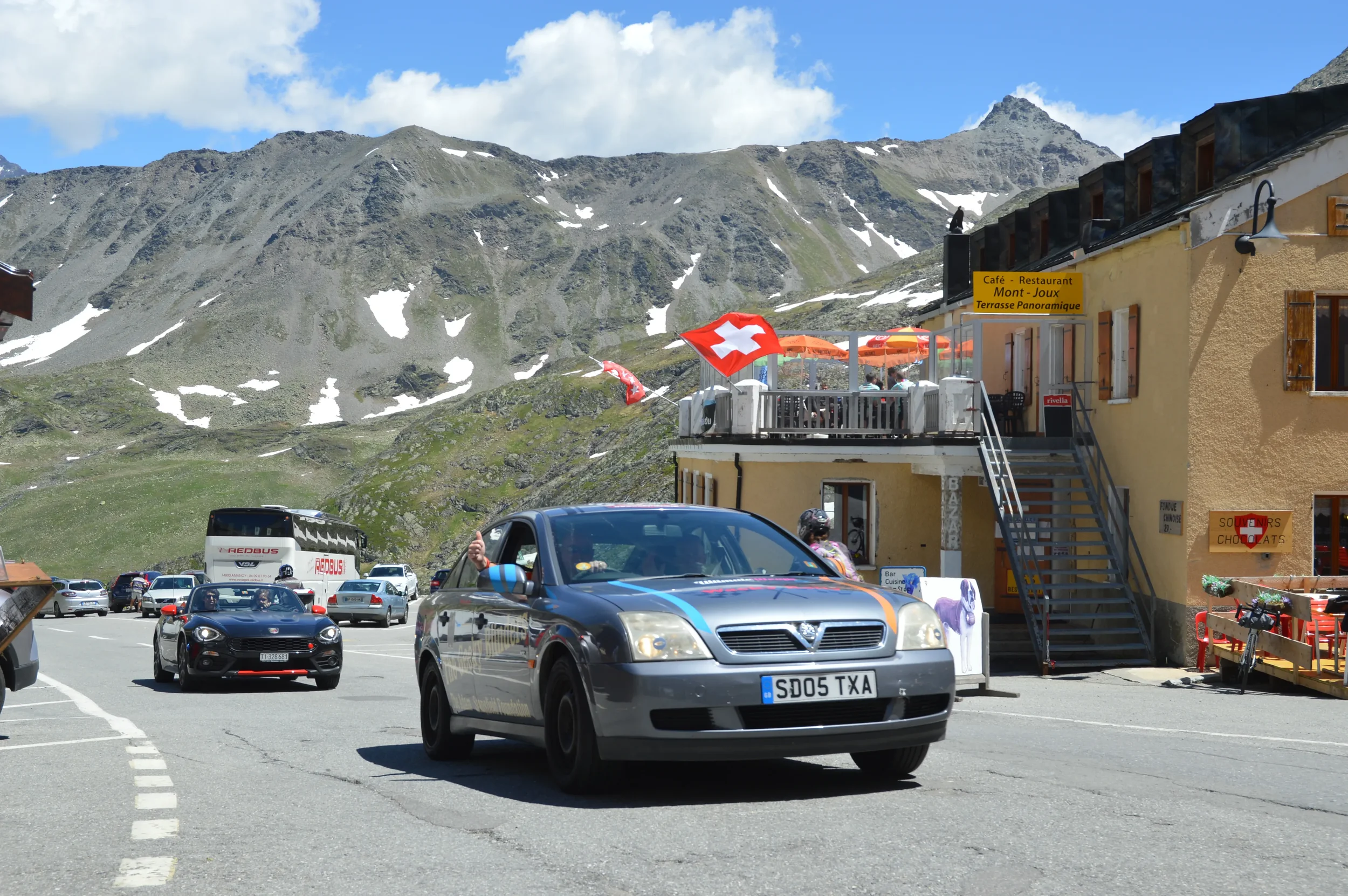 A gray car with a license plate reading 'SD05 TXA' drives through a mountainous area with a cafe and outdoor seating area on the right, decorated with Swiss flags. Several other cars and tour buses are parked, and people can be seen near the building