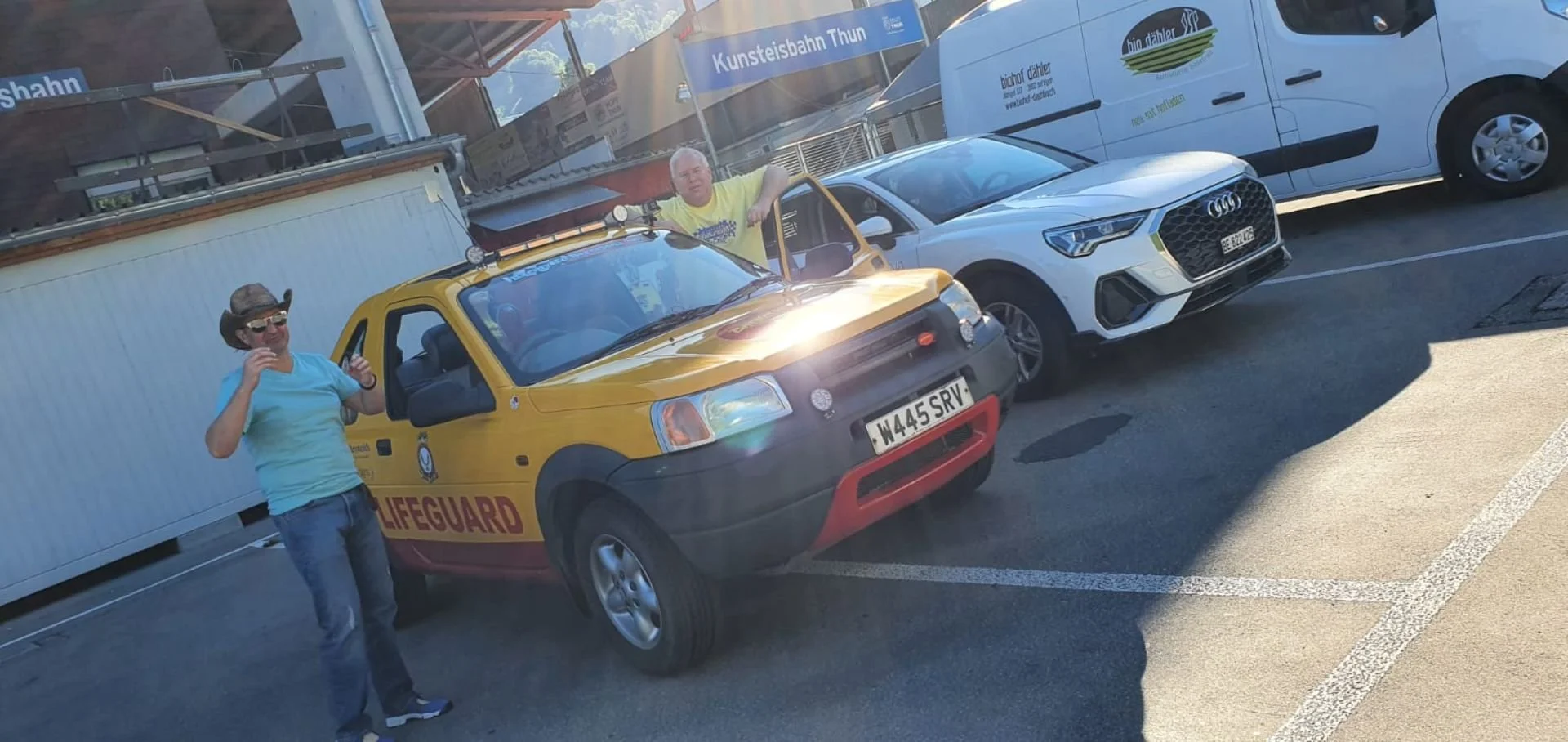 Two men standing in a parking lot, one wearing a cowboy hat and sunglasses giving a thumbs-up, next to a small yellow lifeguard vehicle with a black and red front bumper, and another man sitting inside the vehicle. There are parked cars and a buildin