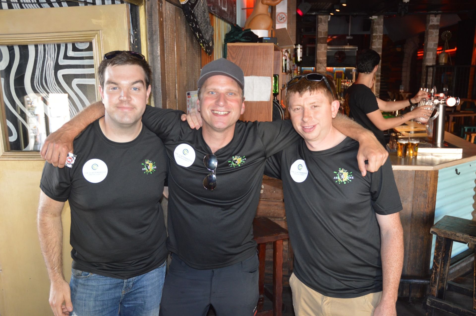 Three men smiling and wearing matching black shirts with badges, standing together with their arms around each other at a bar or restaurant. The background features a bar counter with taps and someone pouring drinks.