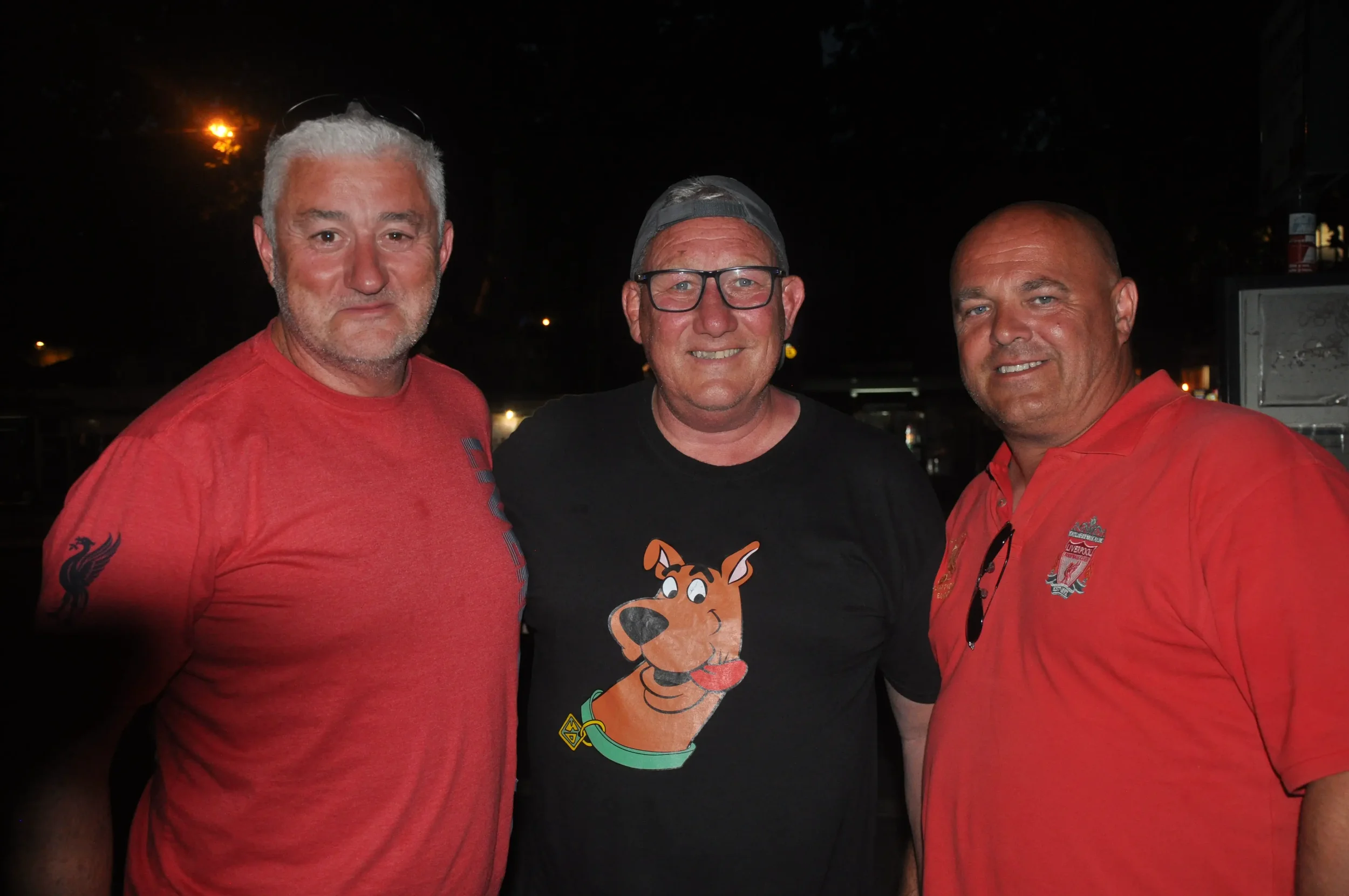 Three men standing closely together, smiling at the camera during an evening outdoor event. The man on the left wears a red T-shirt with a black logo on the sleeve, the man in the middle wears glasses, a backwards baseball cap, and a black T-shirt wi