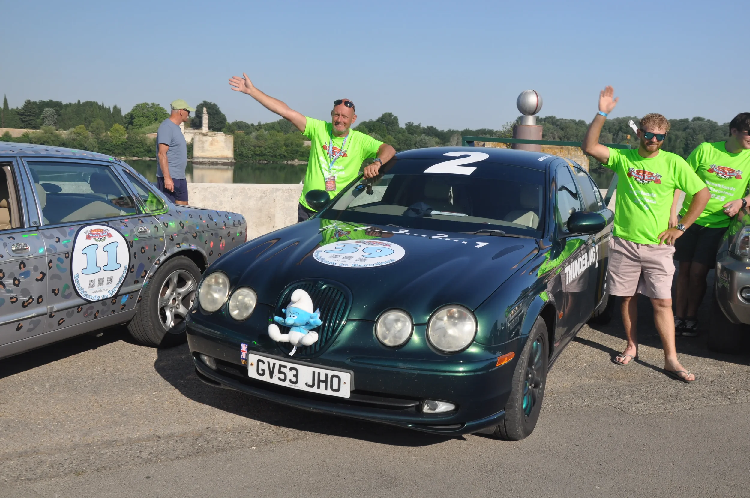 People celebrating near decorated rally cars, with a black car having a number 2 on the windshield and a plush toy hanging on the front, in front of a river and greenery.