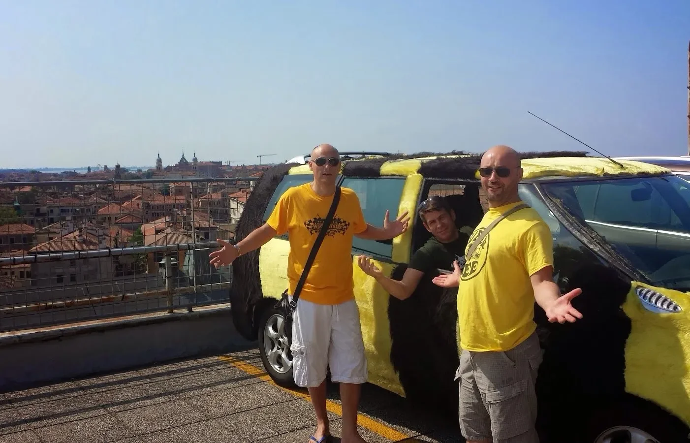 Three men standing in front of a car covered in yellow and black material on a rooftop parking lot, with a city skyline in the background.