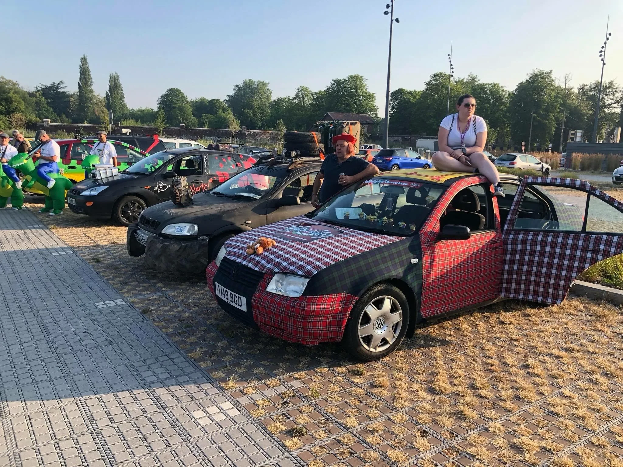 Cars decorated with colorful and themed wrapping and accessories are parked on a paved area next to a grassy field. A woman is sitting on top of a small car covered in plaid fabric, with another man standing nearby. In the background, several people 