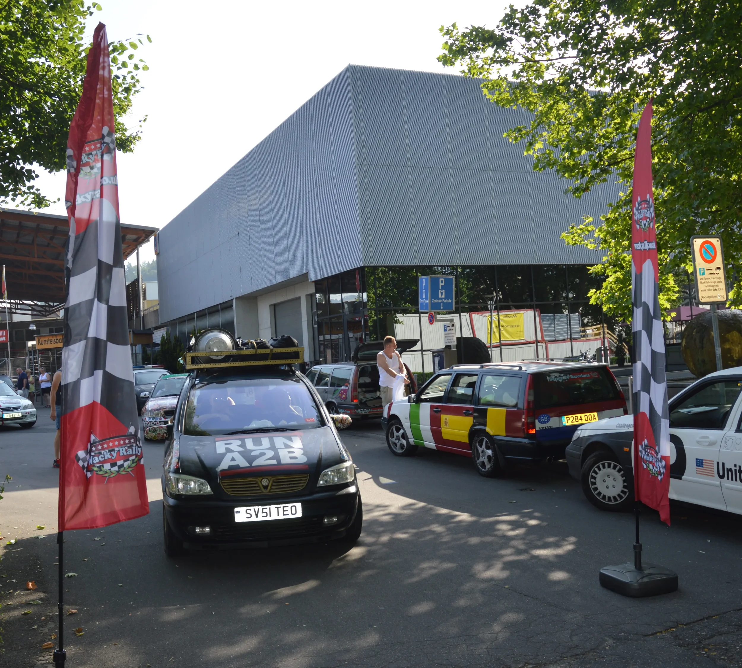 Cars parked in a lot with flags, people, and a modern building in the background.