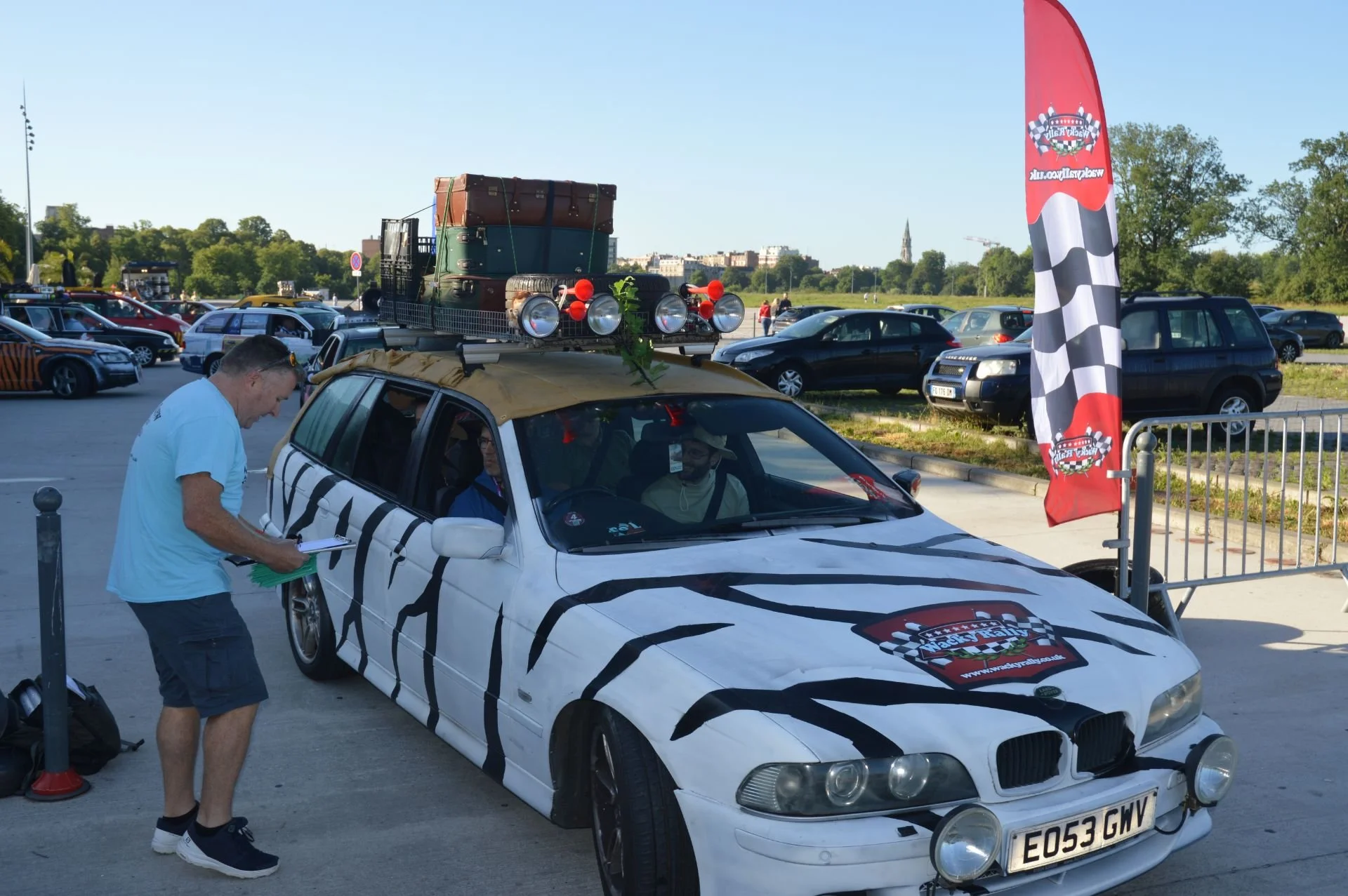 A white BMW with zebra stripes and rally lights on the front, parked outdoors with a roof rack holding vintage trunks and luggage. A man in a light blue shirt is writing or reading something near the car. A red flag with checkered pattern and logo is