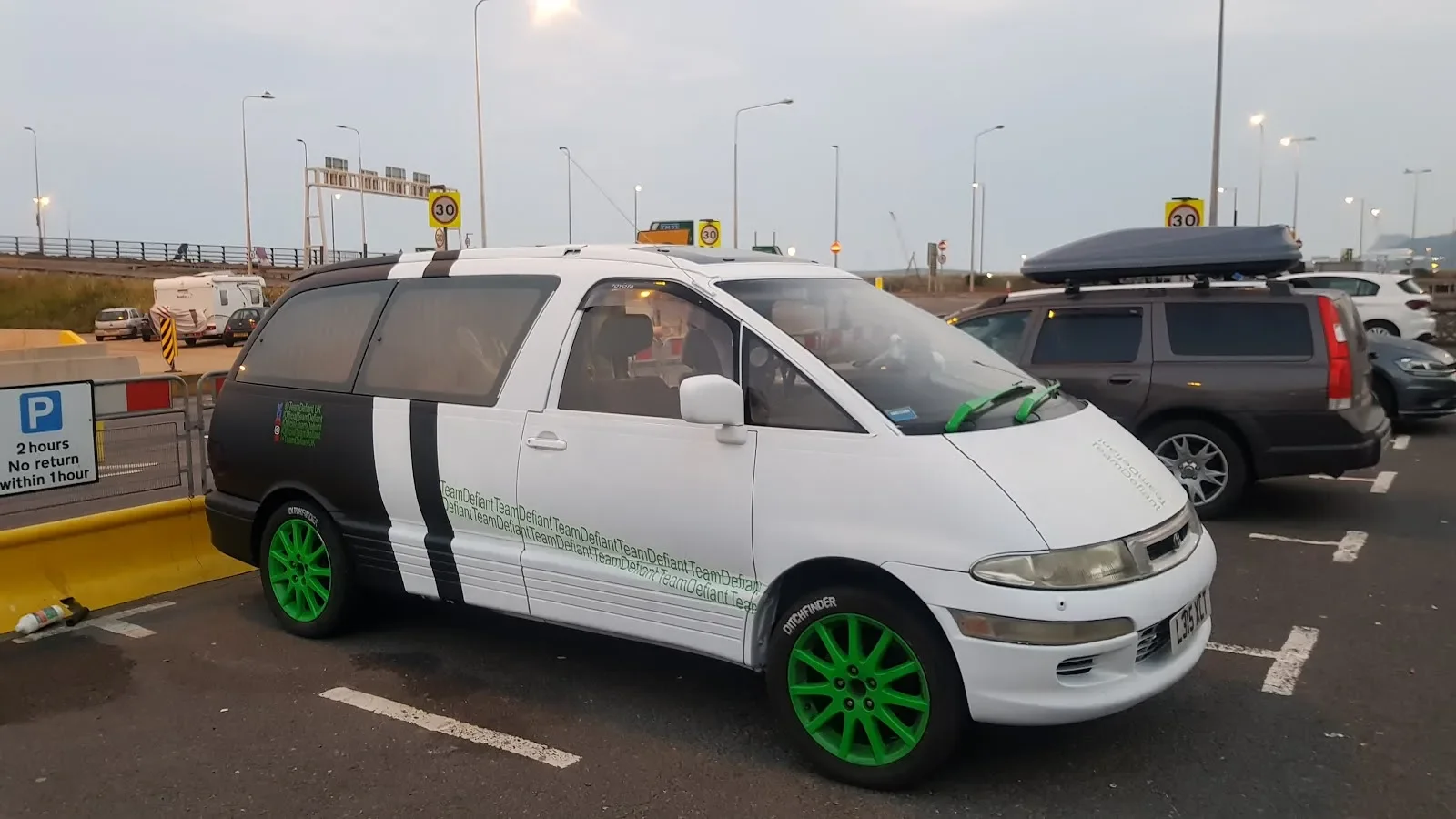 A white van with large green wheels, parked in a parking lot near a yellow barrier. The van has black and white graphics and text along the side, with a small green decal on the driver’s door. Other cars are parked nearby and there are signs indicati