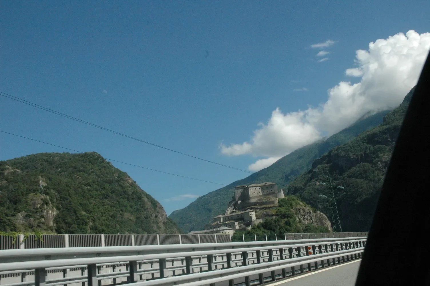 A view of a castle on a hill surrounded by green mountains, seen from a moving vehicle on a highway with guardrails, under a blue sky with some clouds.