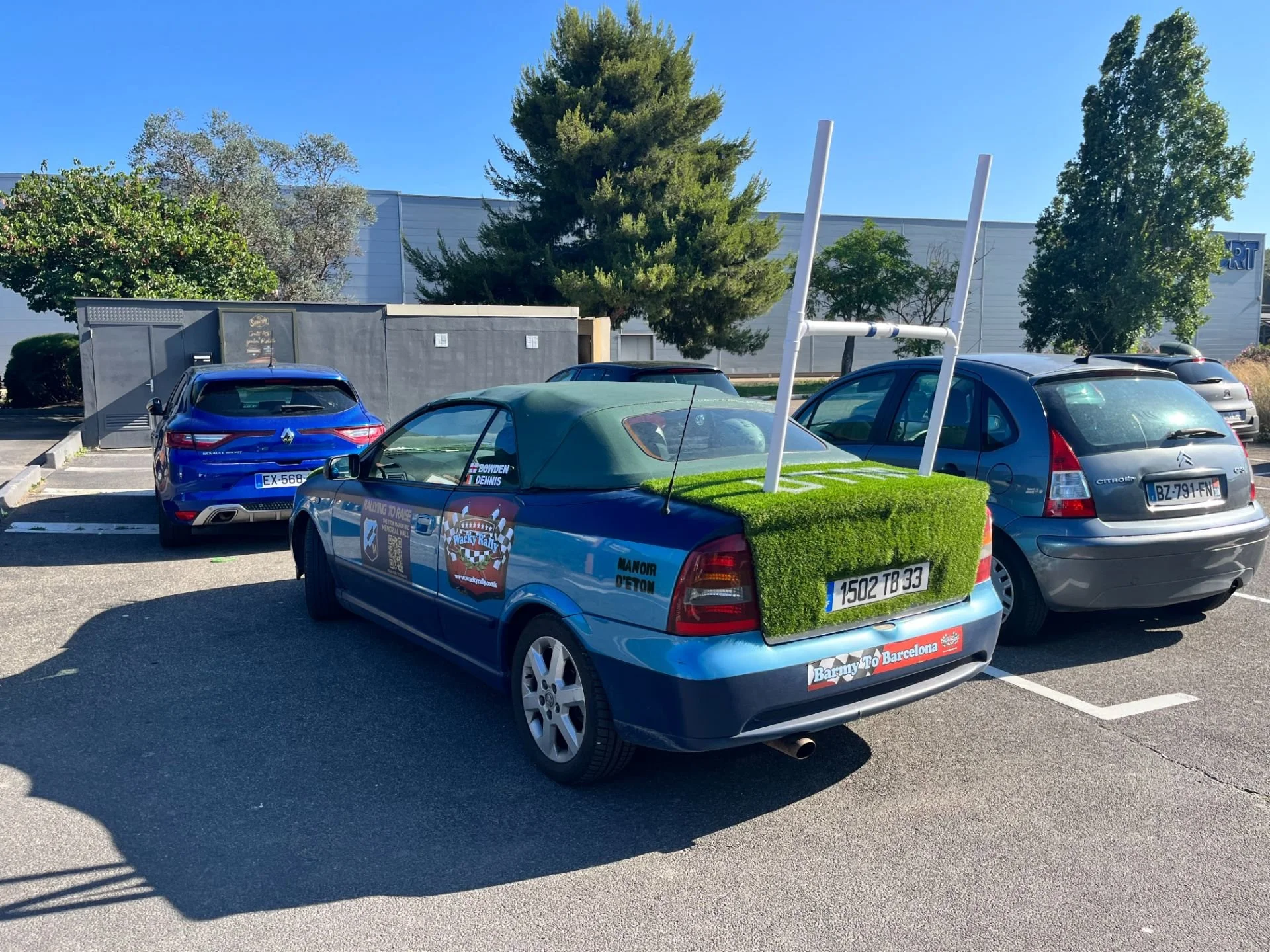 A parking lot with three cars, including a blue convertible with grass and antenna on the back, a silver hatchback, and a blue sedan, under a clear blue sky.