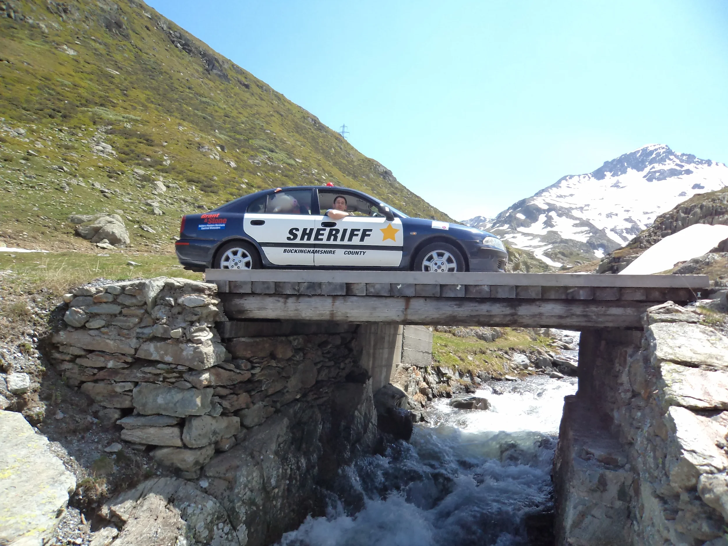 A police car with 'SHERIFF BUCKINGHAMSHIRE COUNTY' written on the side, parked on a small wooden bridge over a stream in a mountainous area.
