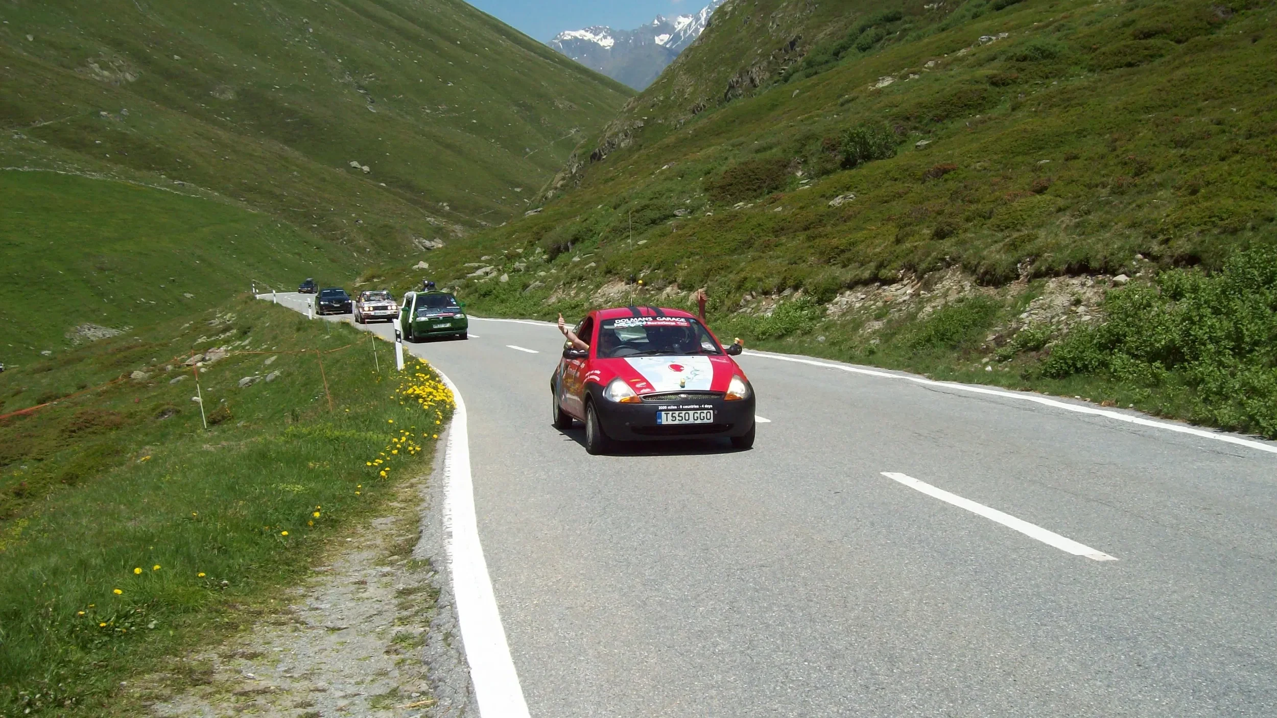 A winding mountain road with a red car in front, with other cars parked along the side, surrounded by green grassy hills and mountains in the distance.