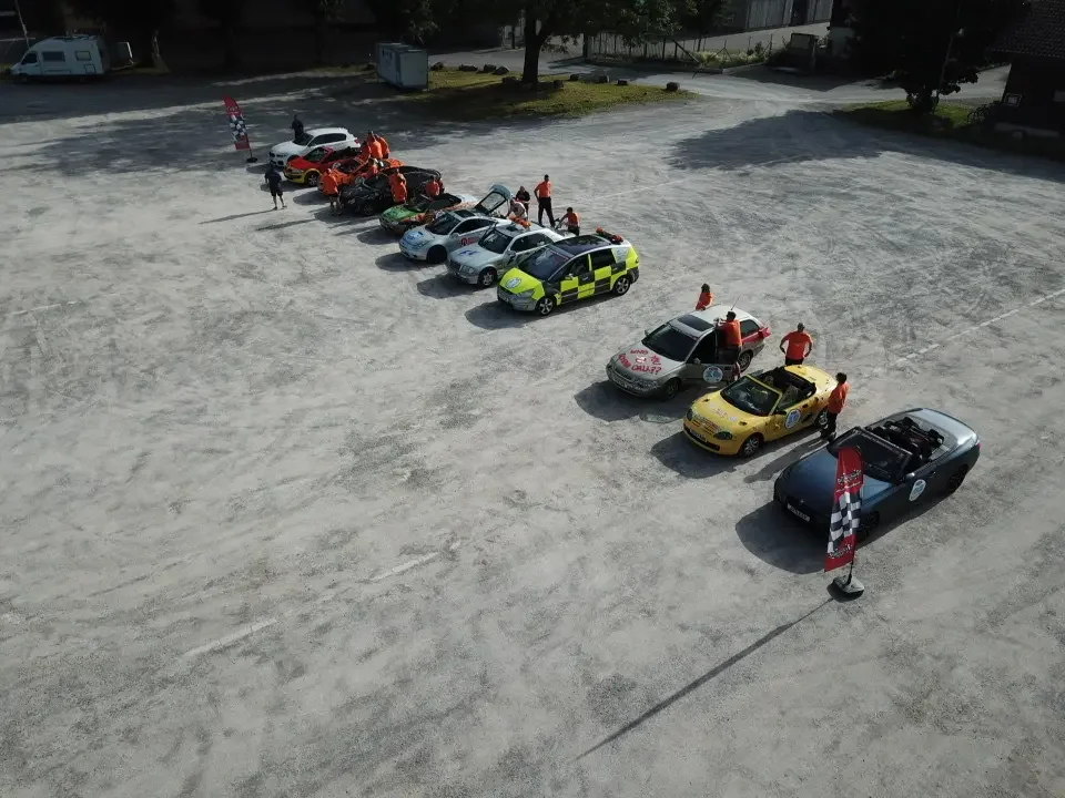 Line of various brightly colored cars, some with racing or emergency markings, parked on a gravel lot with people standing near or around them, and flags or banners nearby.