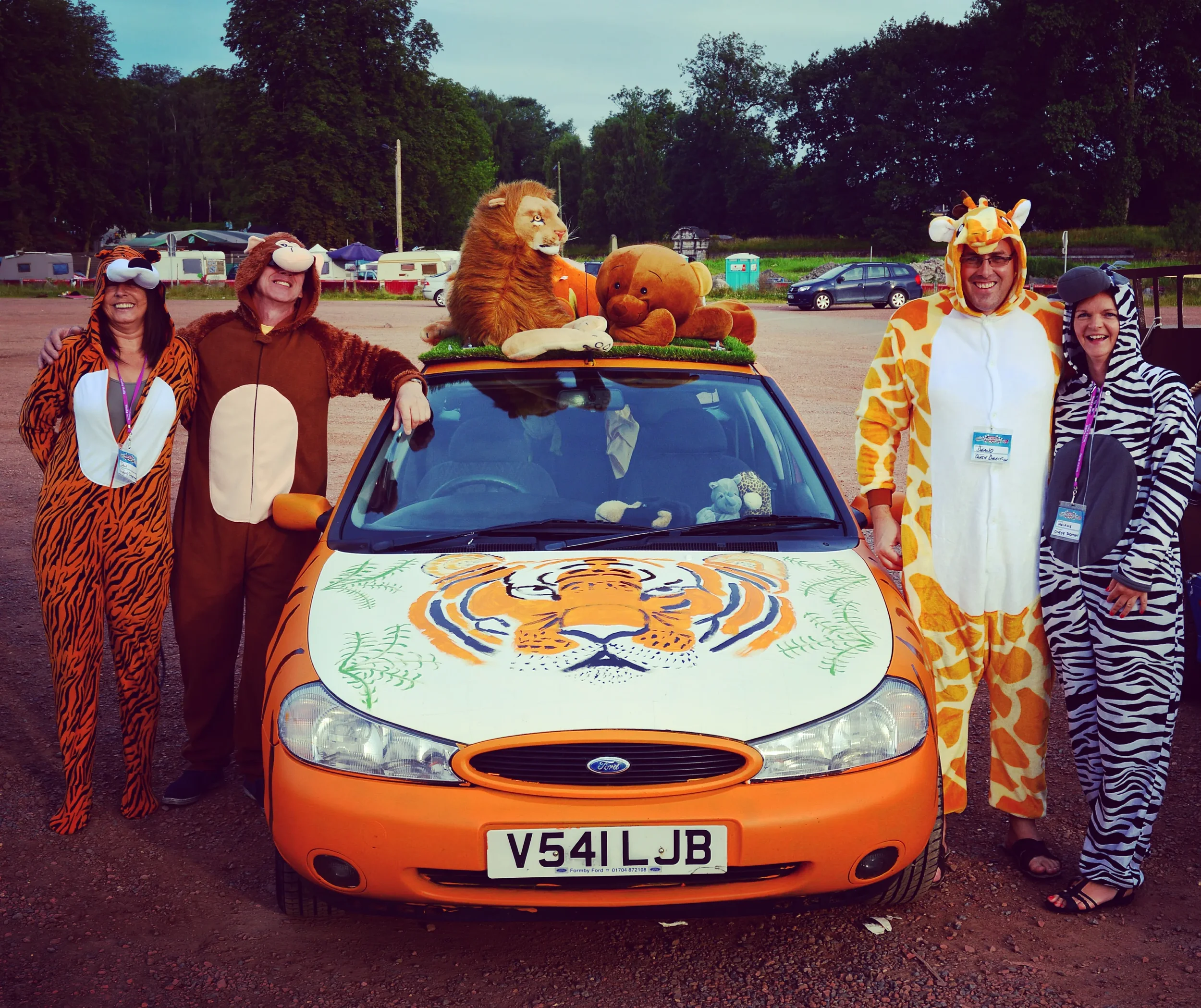 People dressed in animal costumes standing next to a decorated car with a lion, bear, and tiger-themed decorations at an outdoor event.