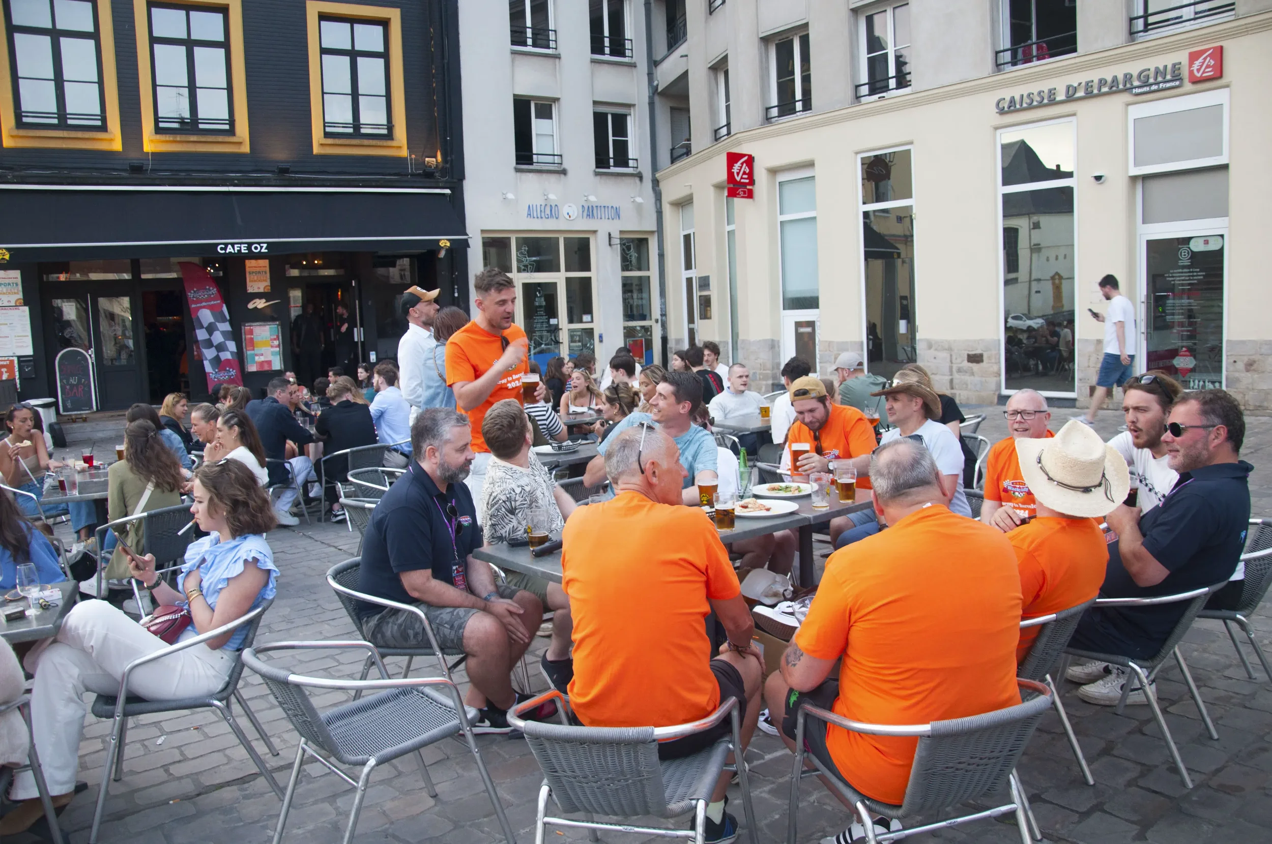 People dining and socializing at outdoor tables on a city street, with some individuals wearing orange shirts and one person standing and speaking.