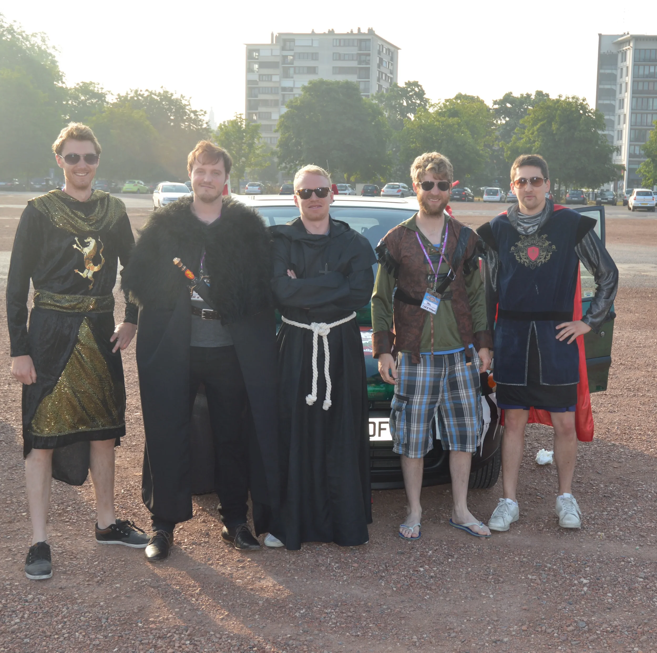 Group of five men dressed in medieval and fantasy costumes standing in front of a black car in an open parking lot with modern buildings in the background during daytime.