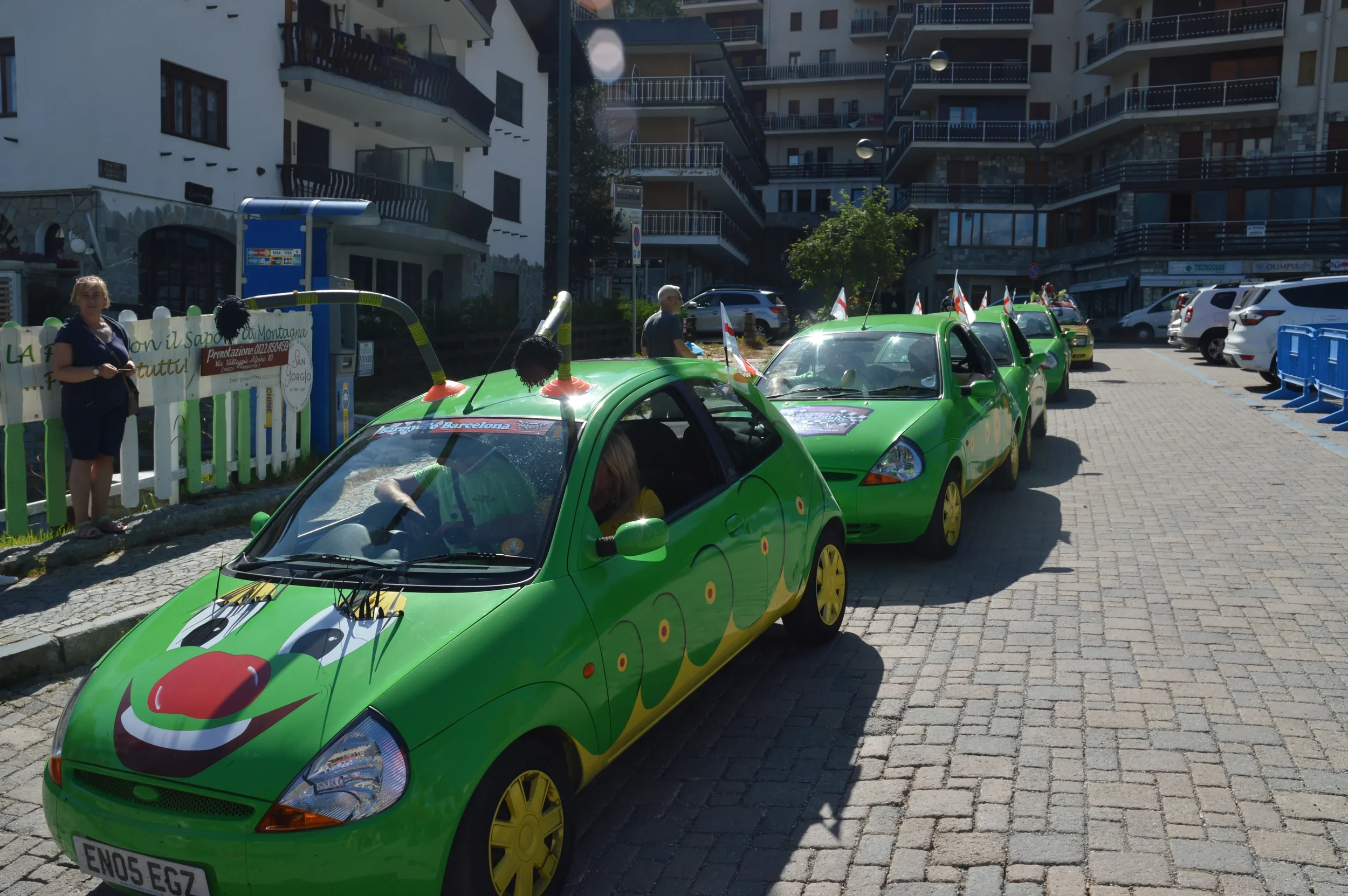 A small parade of green cartoon-themed cars decorated with face designs and antennas is parked on a street in an urban area, with people standing nearby.