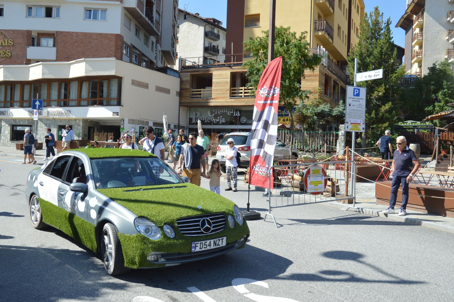 A Mercedes-Benz car covered in artificial grass parked on a city street with pedestrians walking by and a construction site at the corner.
