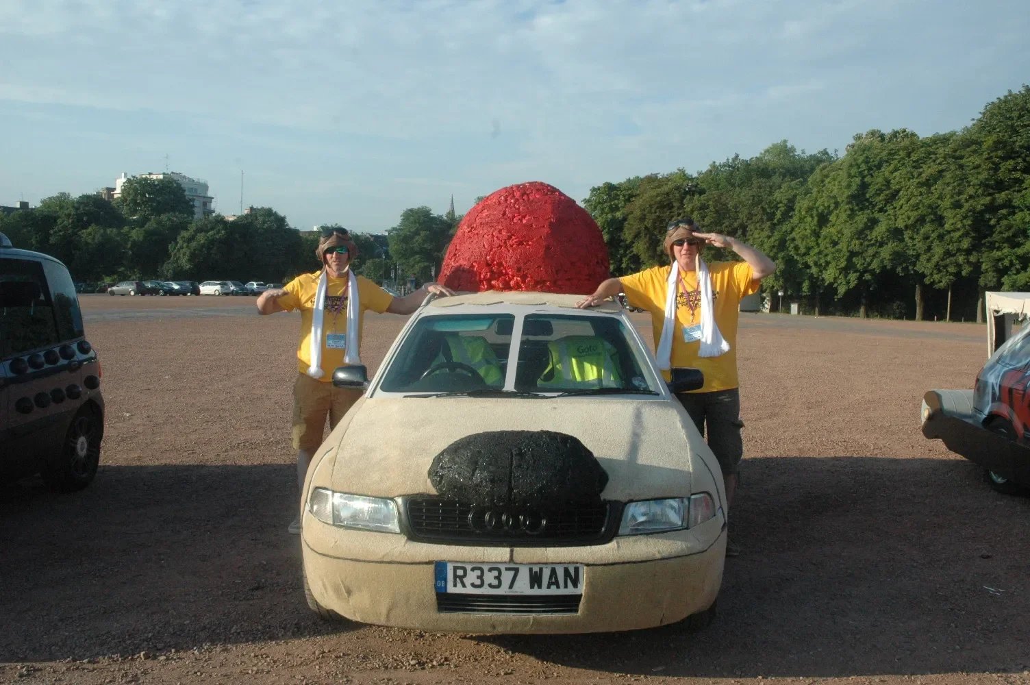 Two women standing next to an old beige Audi car with a large red and black sculptural object on its roof, in a parking lot during the daytime. The women are wearing yellow shirts, sunglasses, and white scarves, and are saluting.