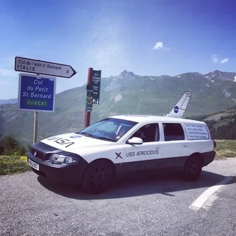 A white vehicle with 'USS Atrocious' written on the side, parked near mountain signs in a mountainous area with clear blue sky.