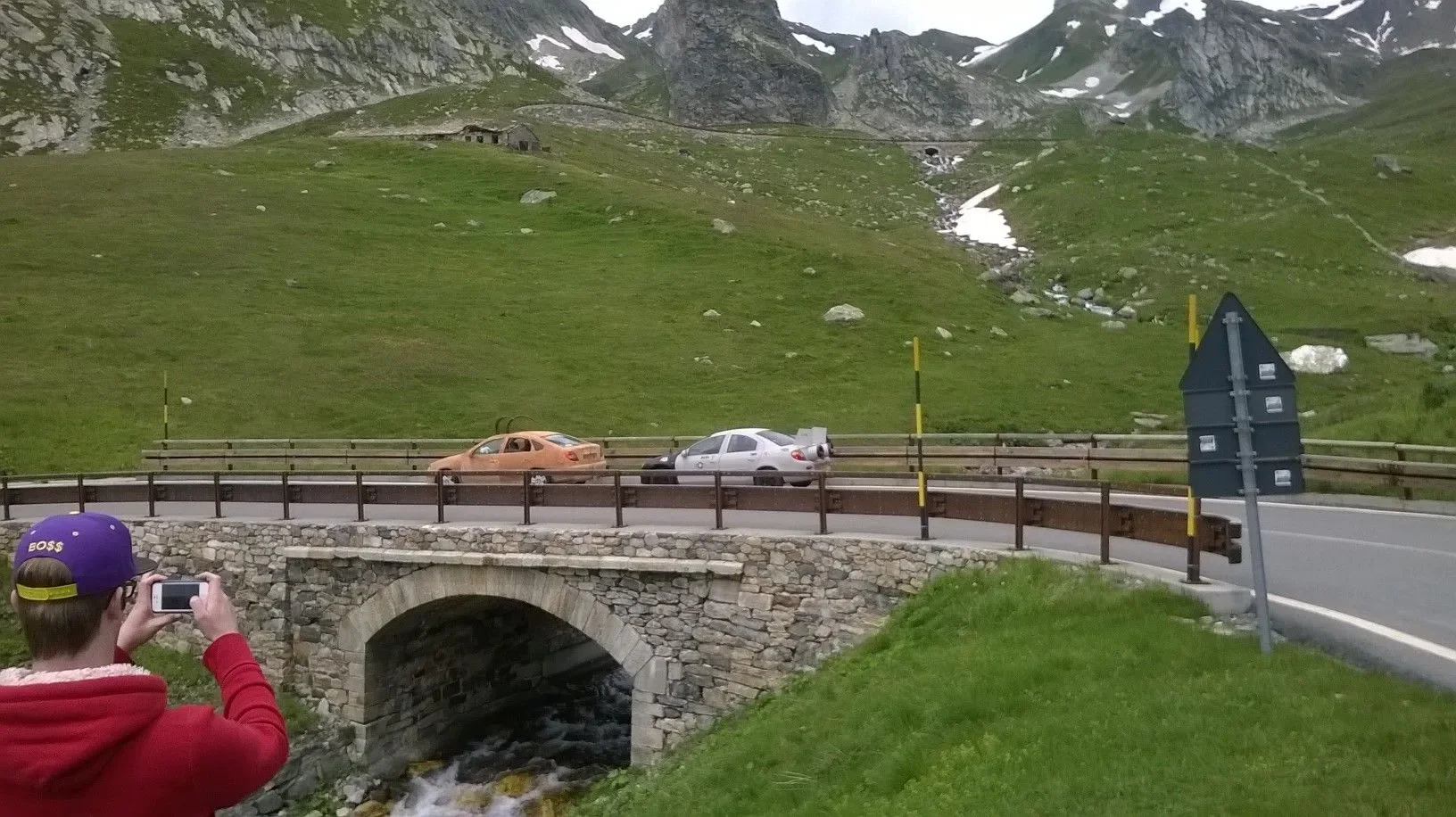 A person wearing a purple baseball cap and red hoodie taking a photo of two cars on a mountain road, with a stone bridge over a small stream in the foreground, and green hills and mountains in the background.