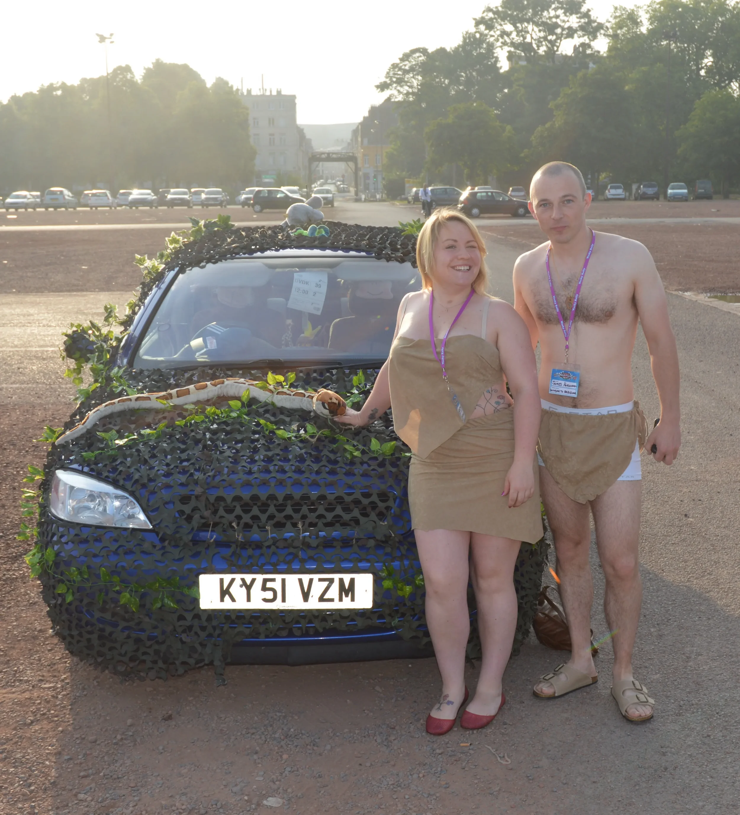 Two people dressed in makeshift medieval costumes standing next to a decorated car in an outdoor parking lot during daytime.