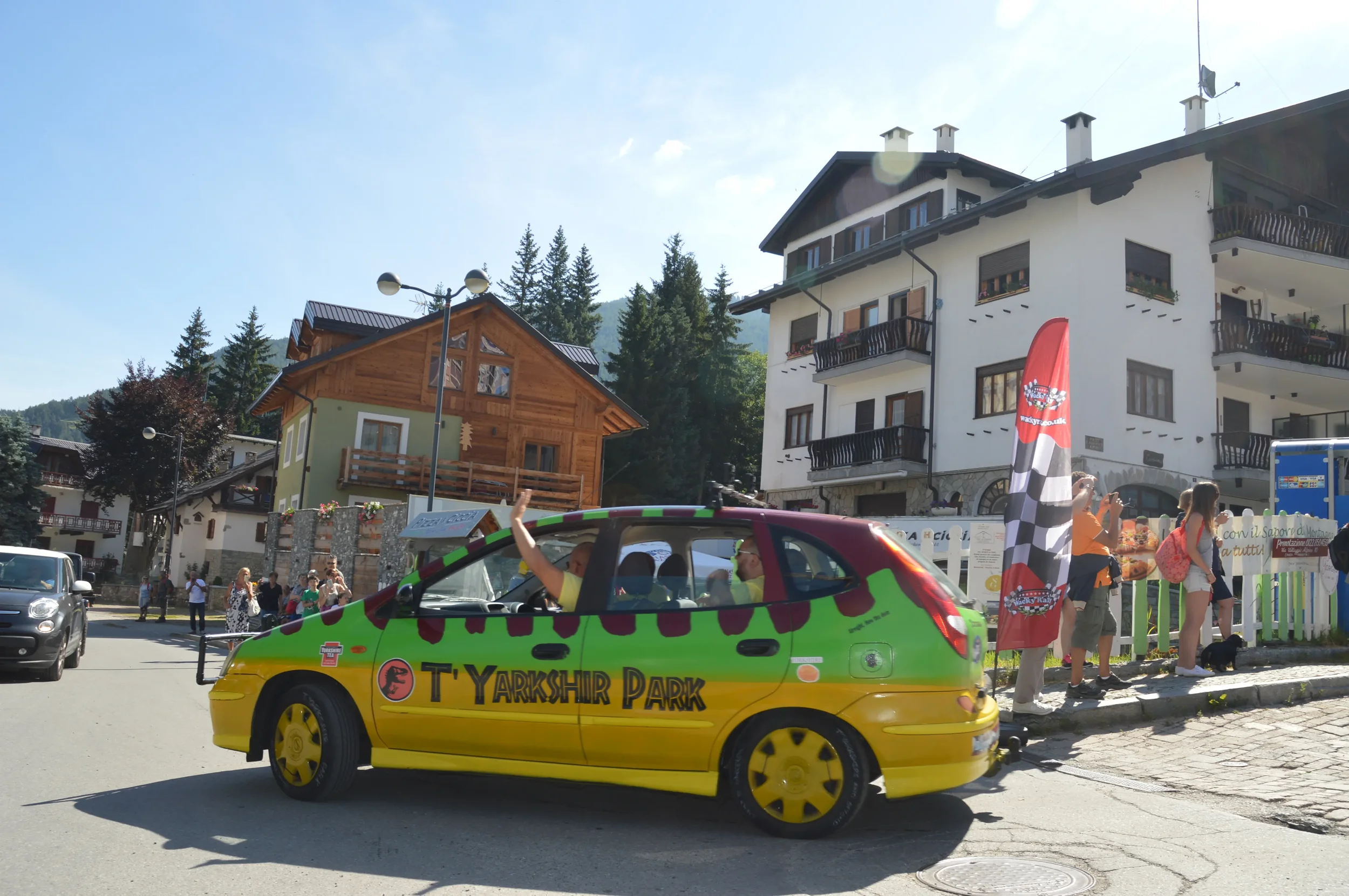A colorful car with the words 'T. YARKSHIR PARK' on the side, driving past a crowd of people in a mountain town, with buildings and pine trees in the background.