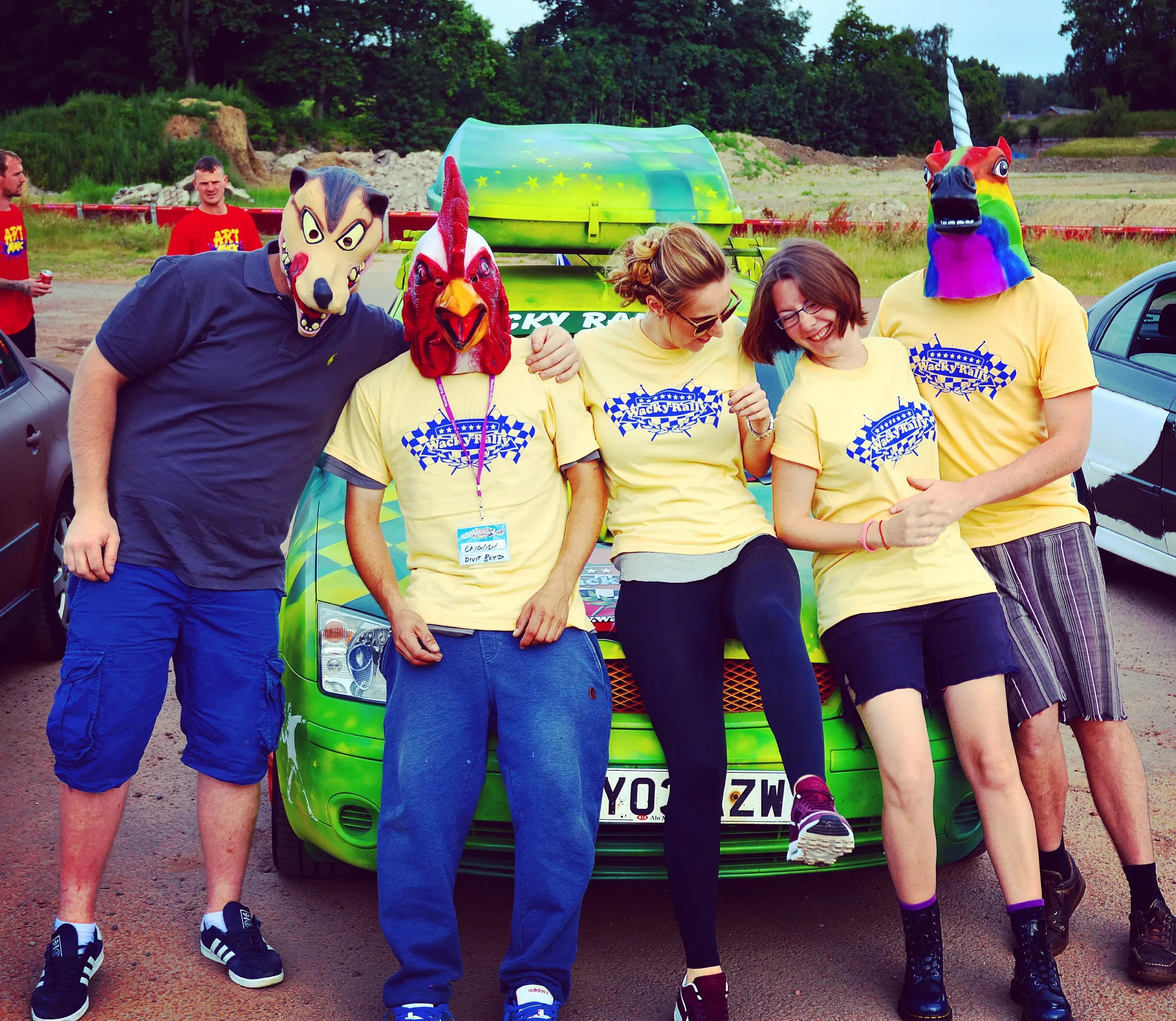 Group of five people wearing masks and yellow T-shirts, standing in front of a green car at an outdoor event with grass and trees in the background.